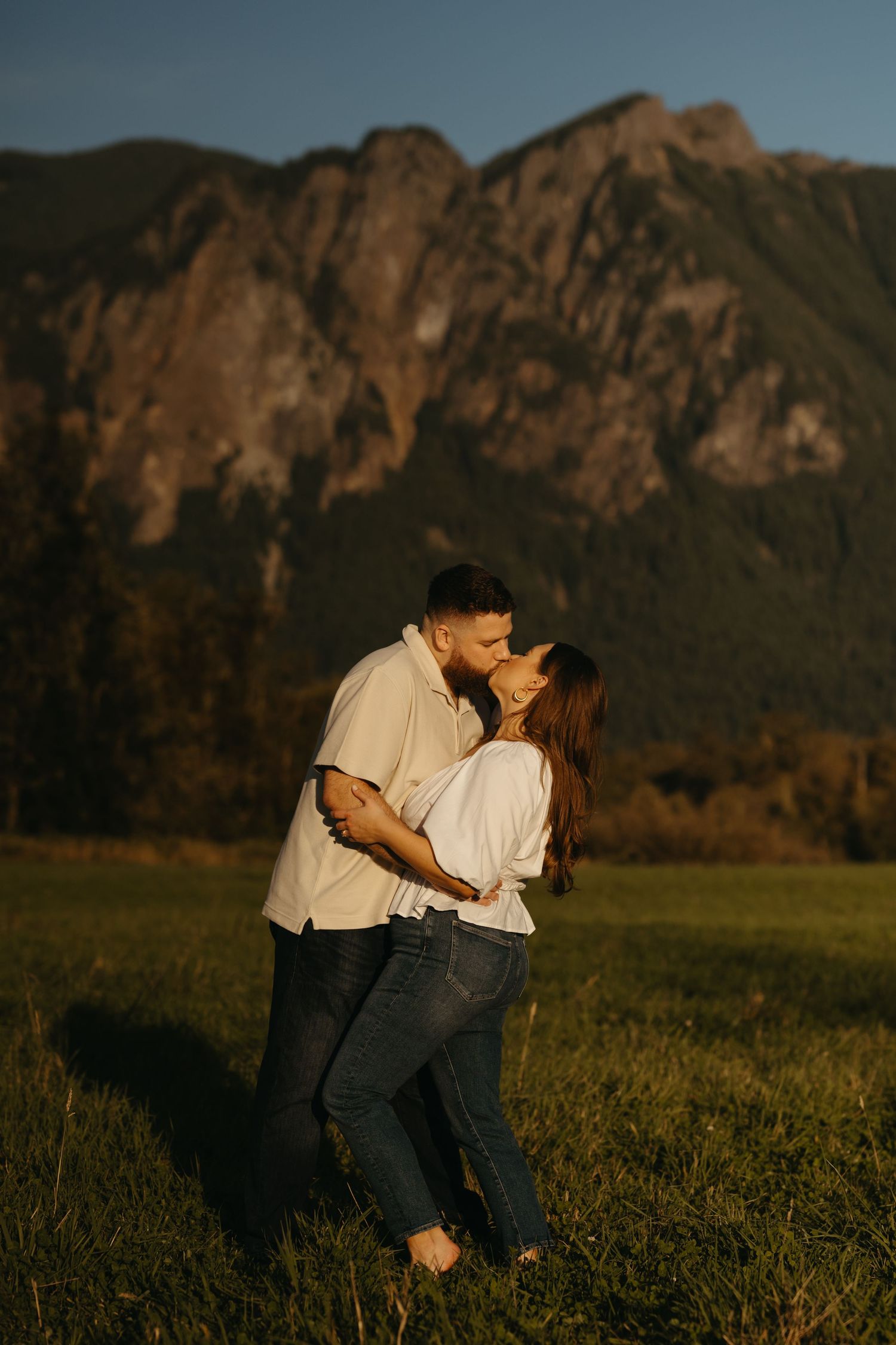 Couple embracing in field with dramatic mountain backdrop during sunset golden hour.