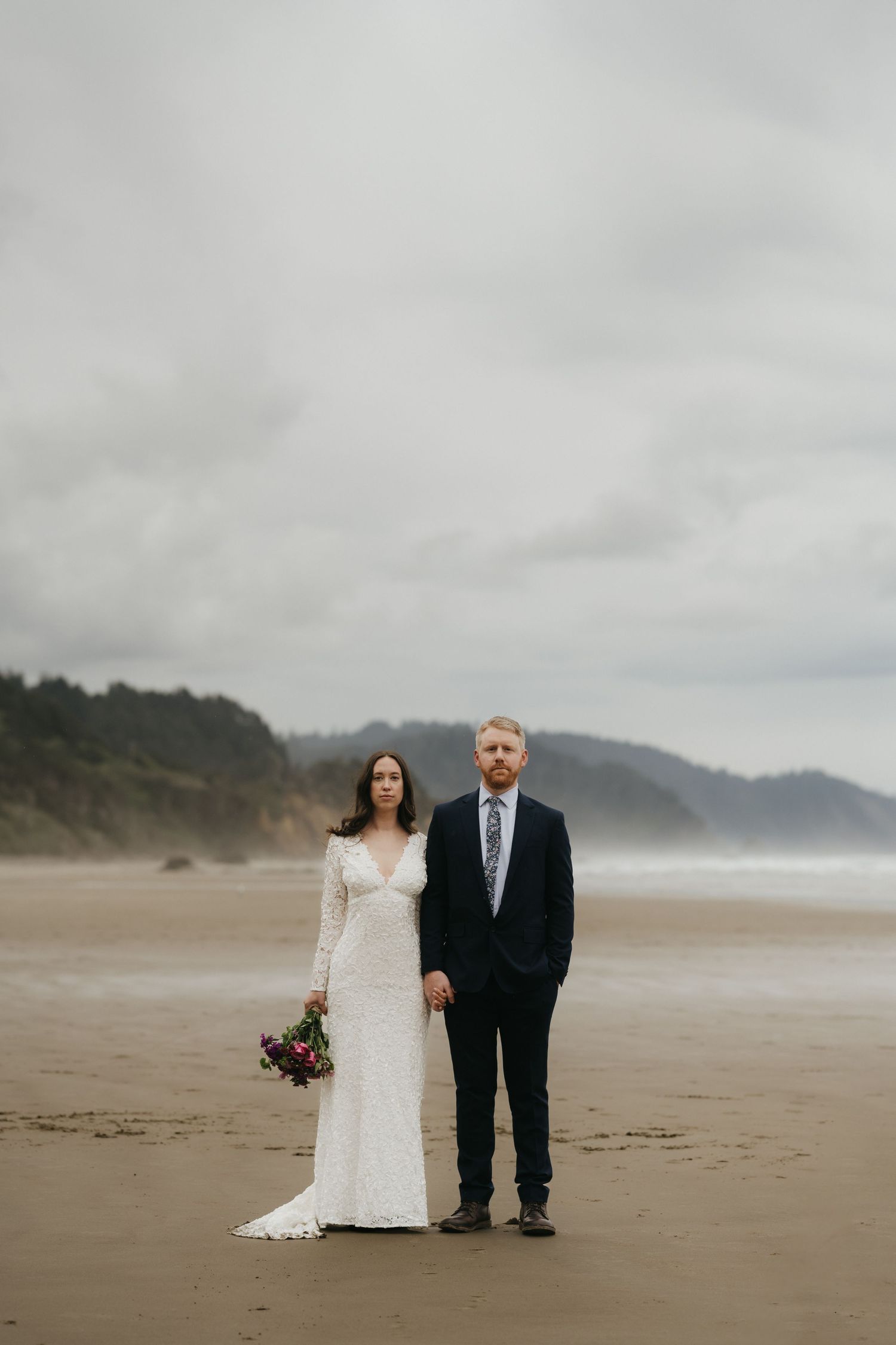 Newlyweds standing together on a moody beach with mountains in the background.