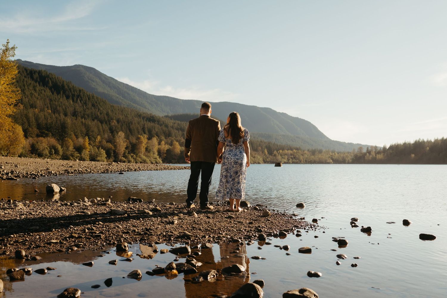 A couple stands together overlooking a serene lake surrounded by mountains and autumn trees.
