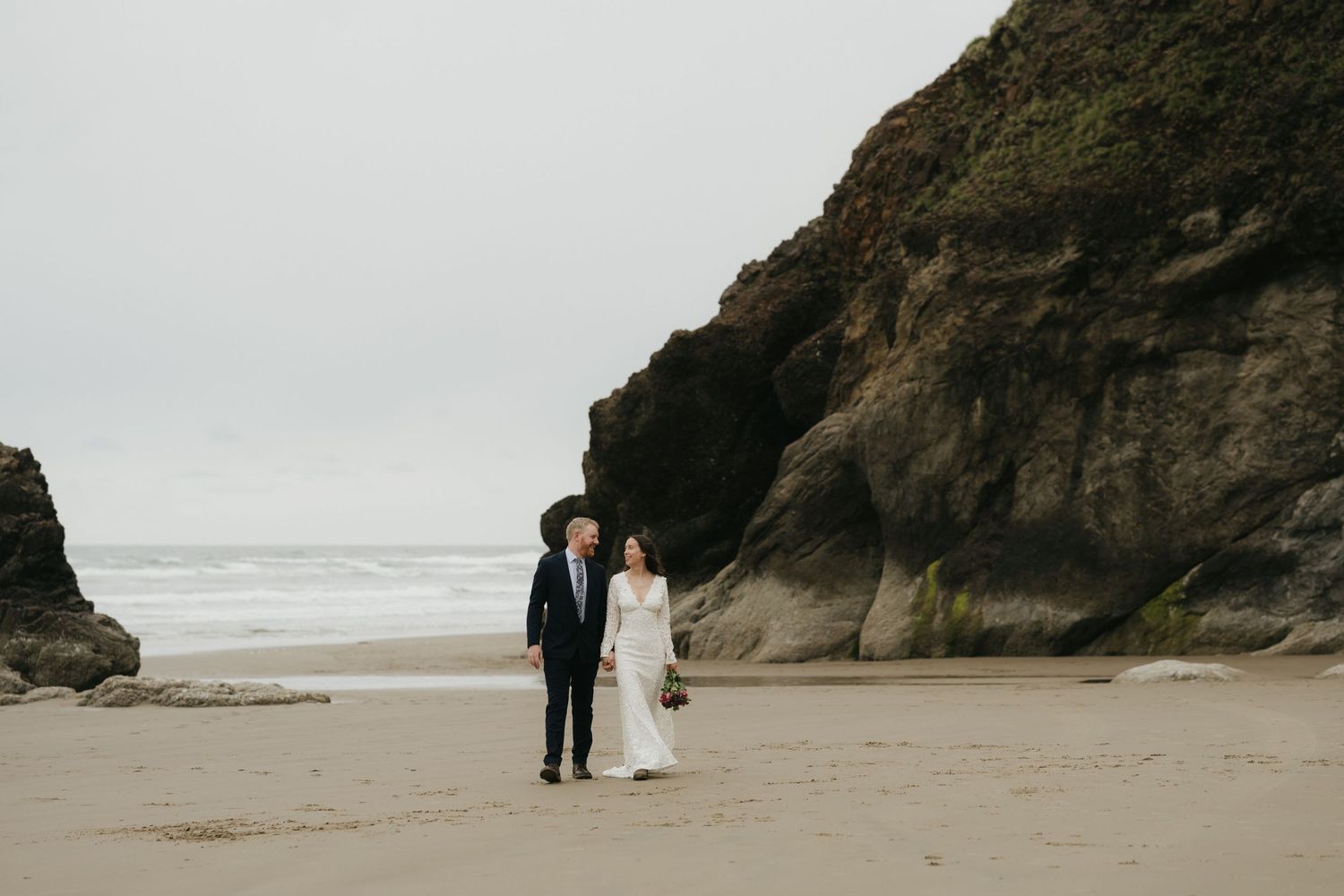 Bride and groom standing together on sandy beach surrounded by towering rock formations.
