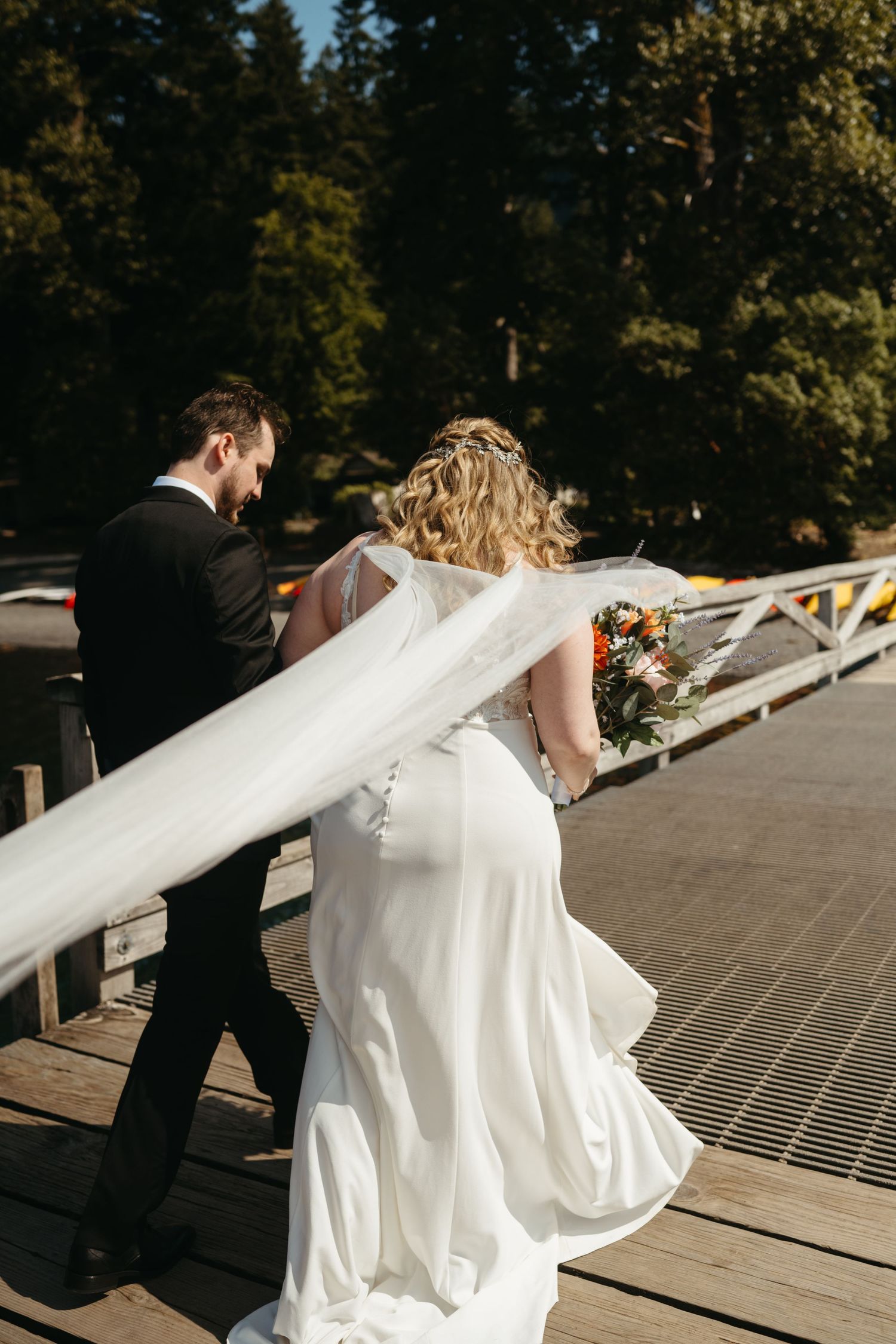 Wedding couple walking on a wooden dock with flowing white dress trailing behind.
