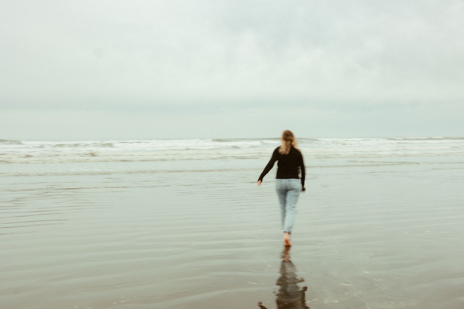 Figure strolling barefoot along the shoreline on a cloudy oceanside morning.