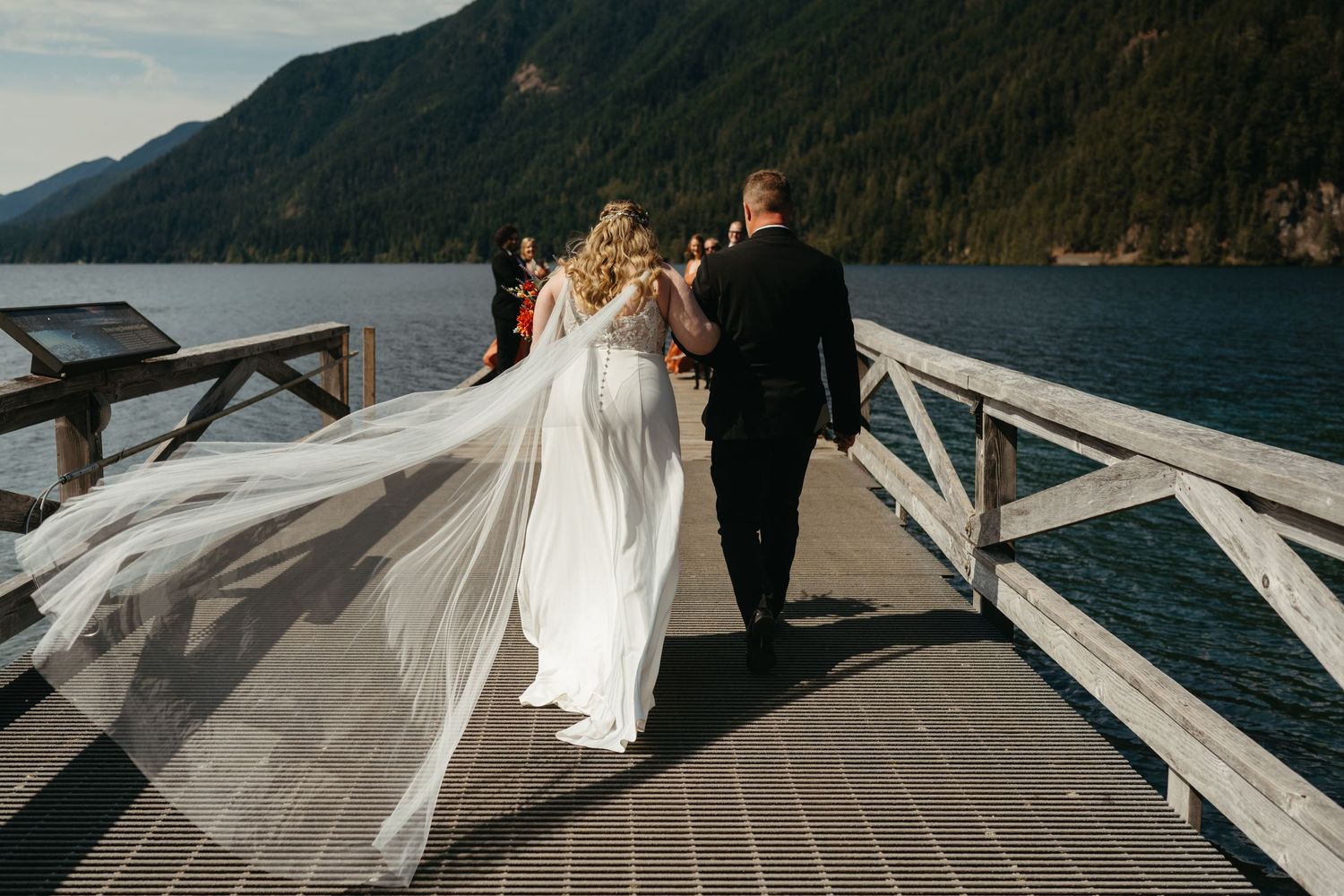 Bride and groom walking on wooden dock with mountain lake views and flowing veil.