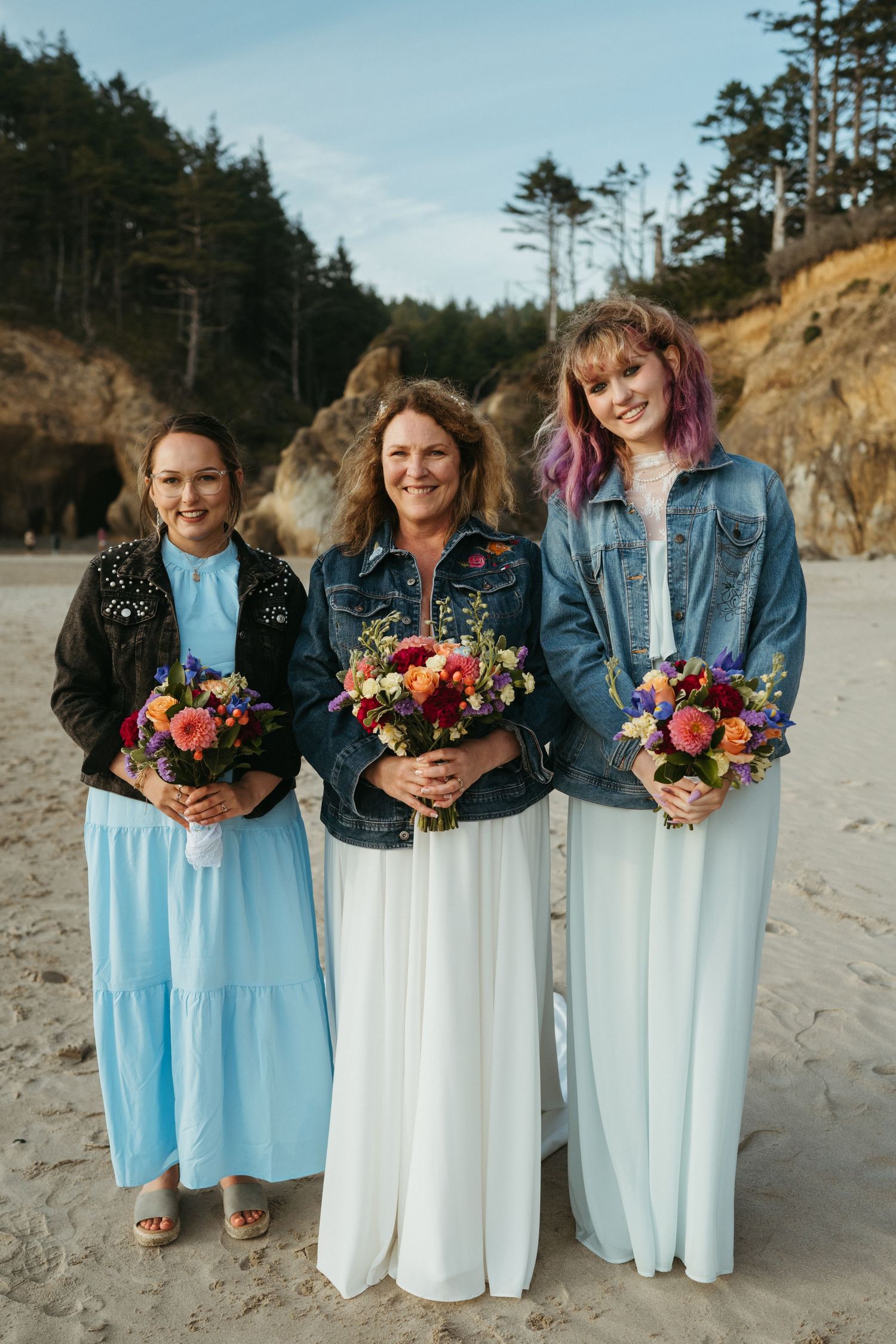 Three bridesmaids in denim jackets and long dresses hold colorful bouquets on a beach.