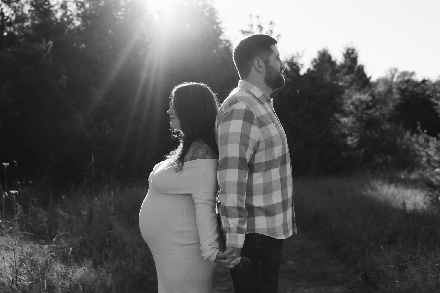 Black and white maternity photo of a couple holding hands in a sunlit field, woman showing baby bump.