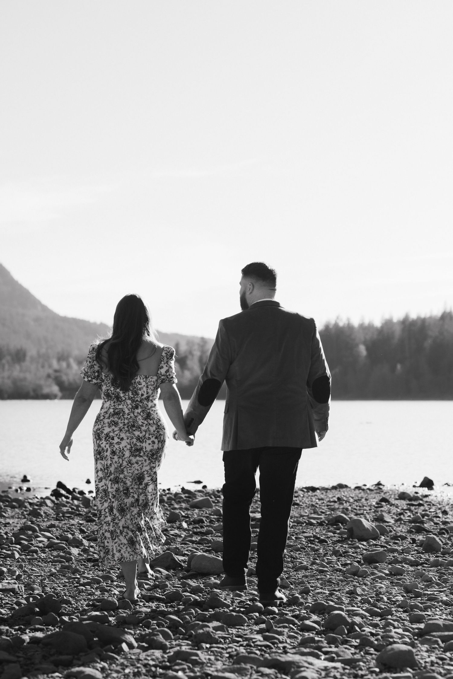 A black and white photo of a couple walking together on a rocky beach with mountains in the distance.