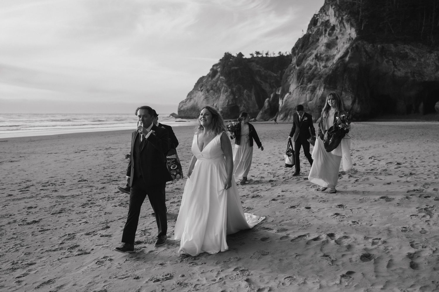 A wedding party walks along a dramatic coastal beach with cliffs in the background.
