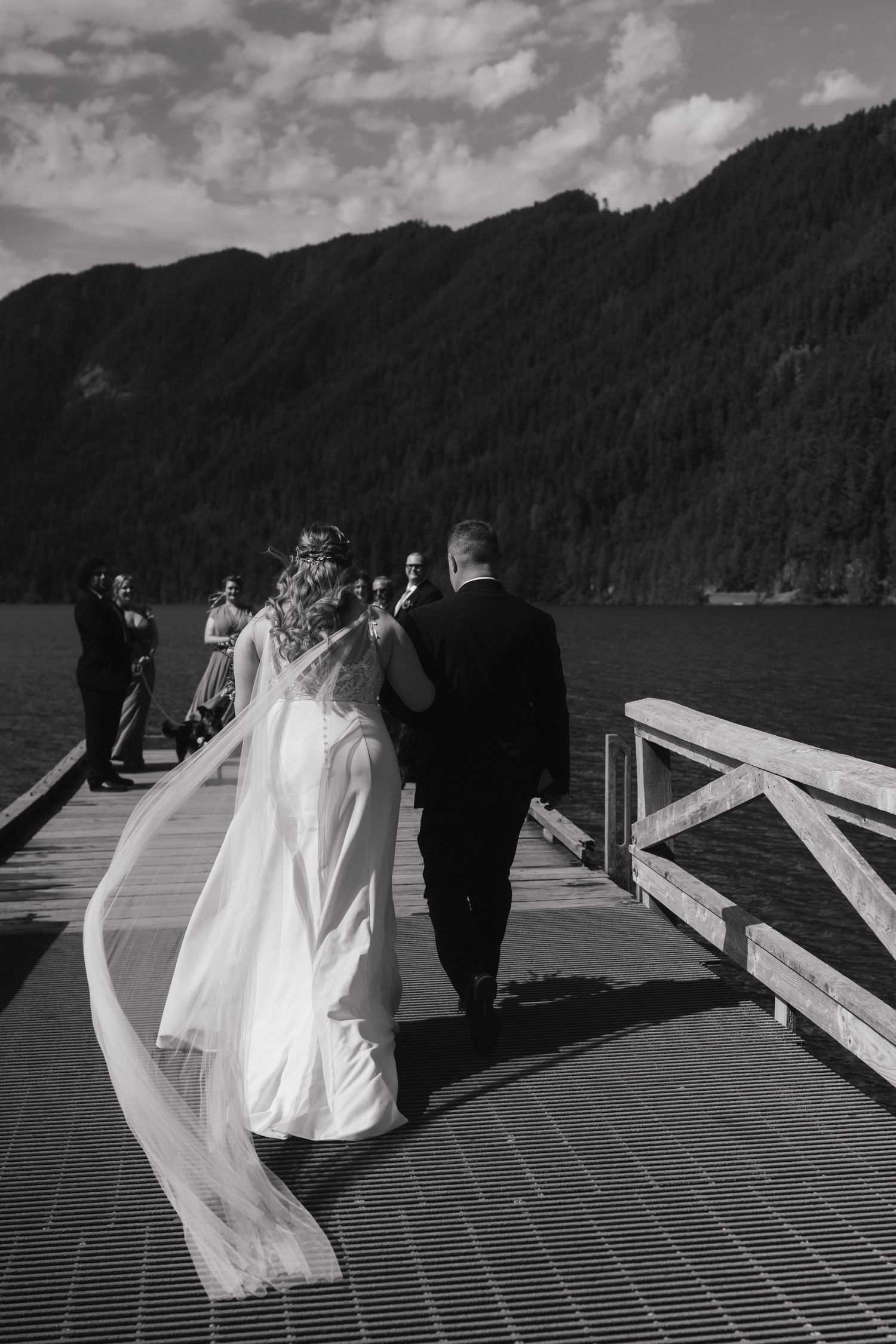Black and white photo of newlyweds walking on lakeside dock with veil flowing.