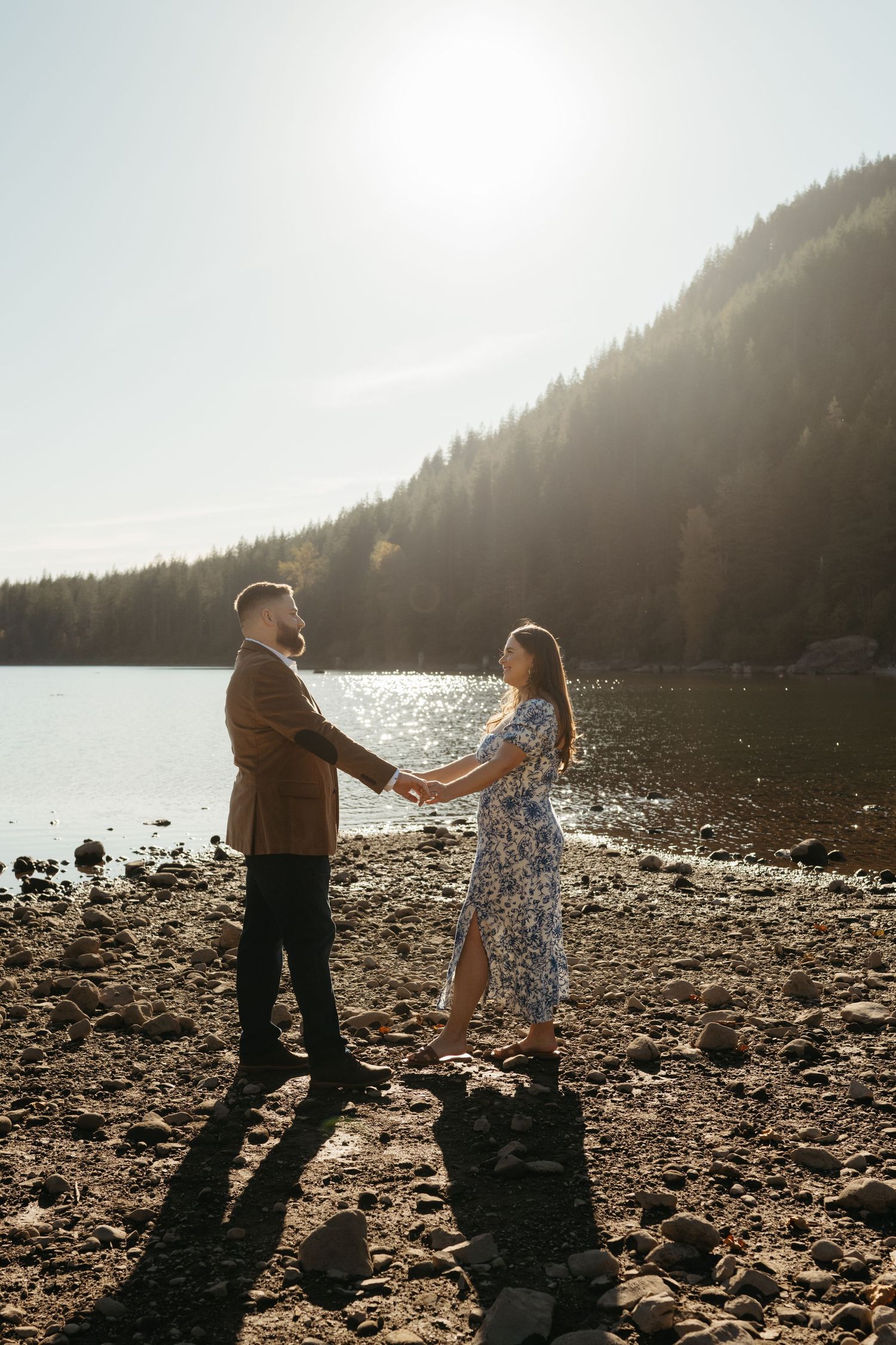 A couple dances on a rocky beach with sunlit mountains and forest in the background.
