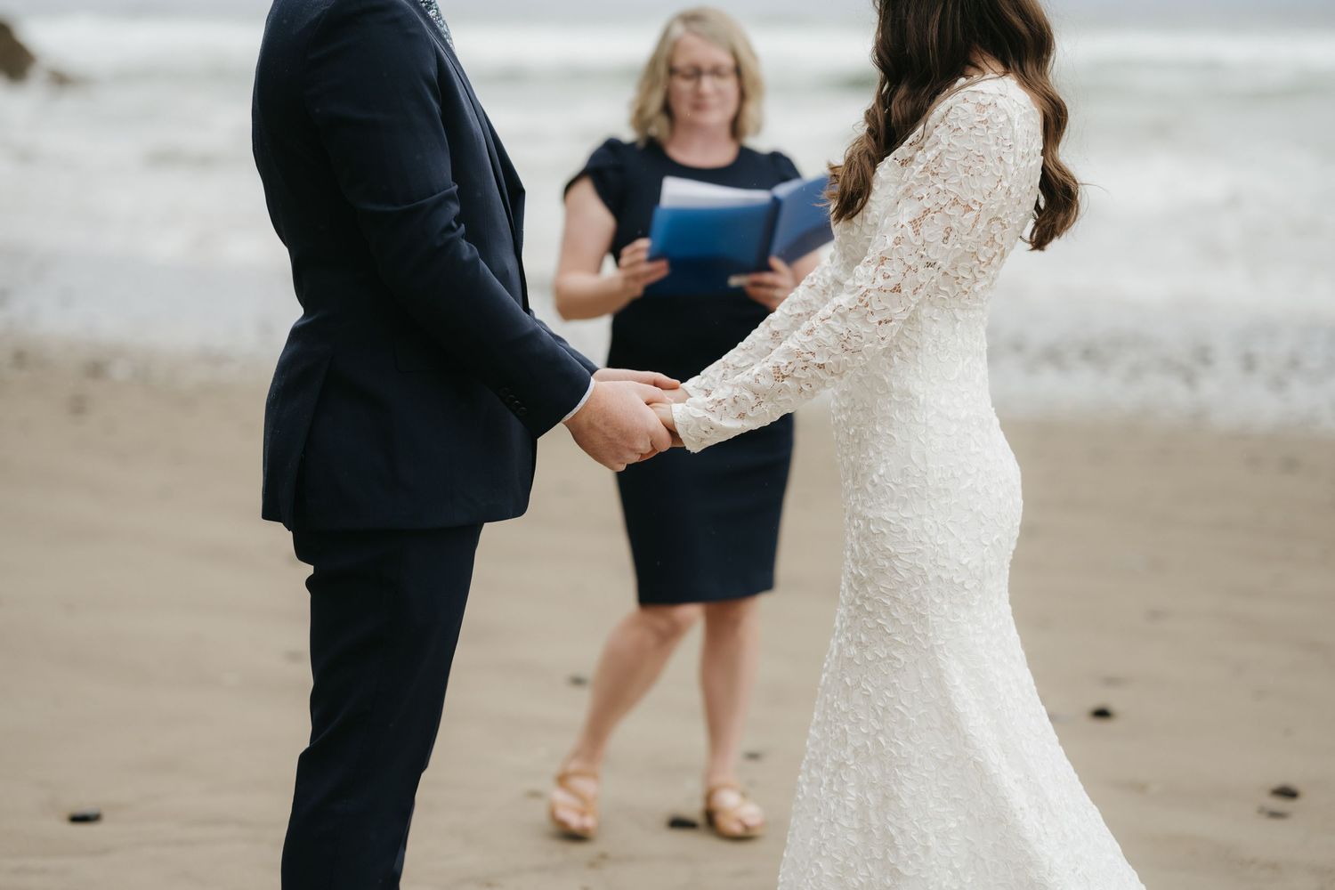 A couple holds hands during their intimate beach wedding ceremony with an officiant in a navy dress.
