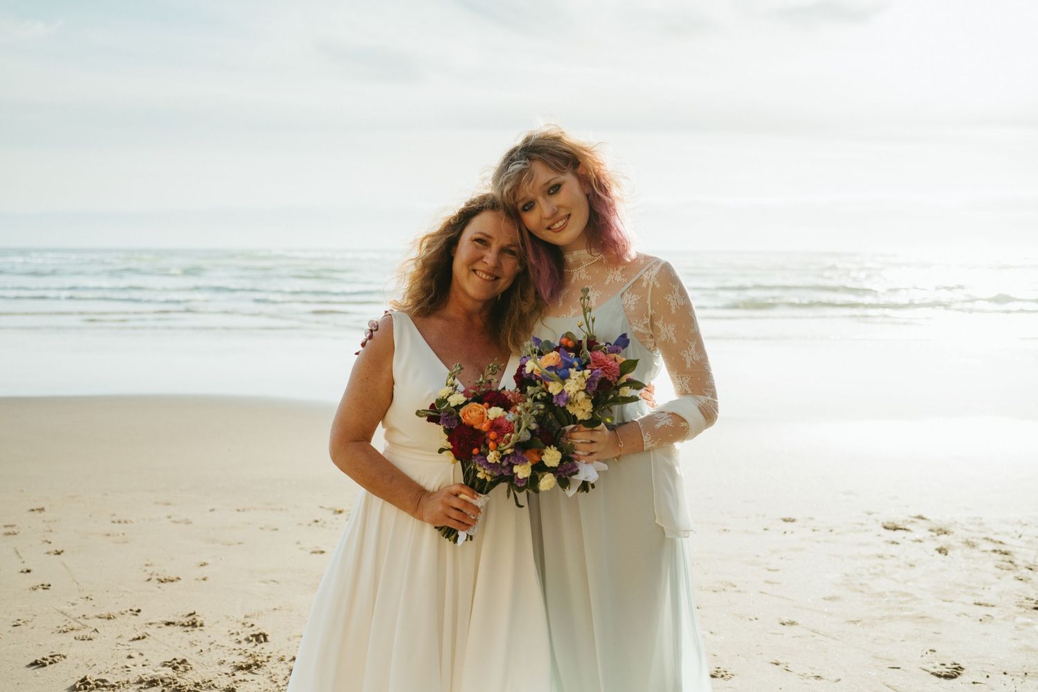 Two attendants with floral bouquets pose together on a sunny beach.