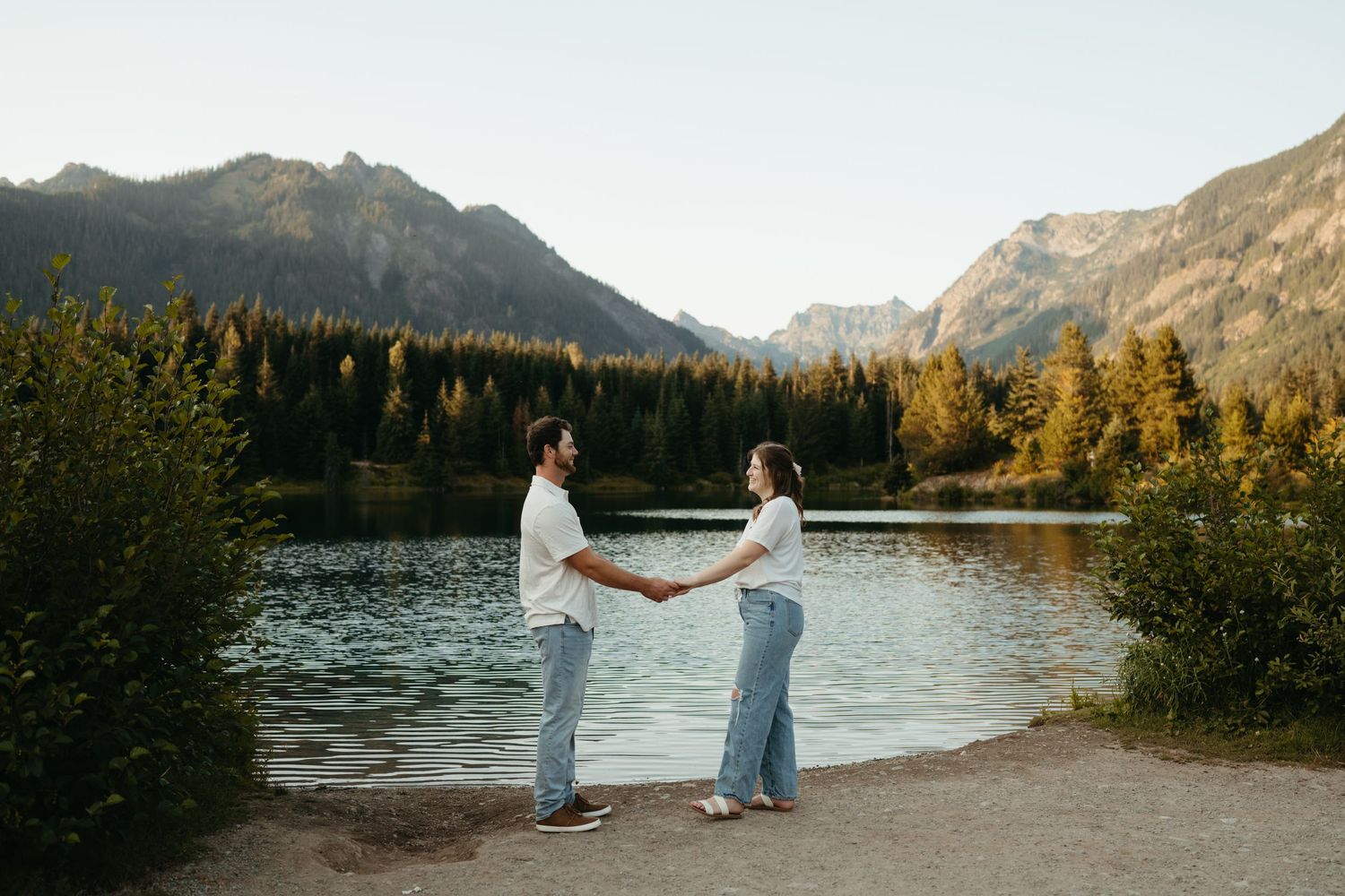 Couple holds hands at sunset by mountain lake with evergreen forest and peaks in background.