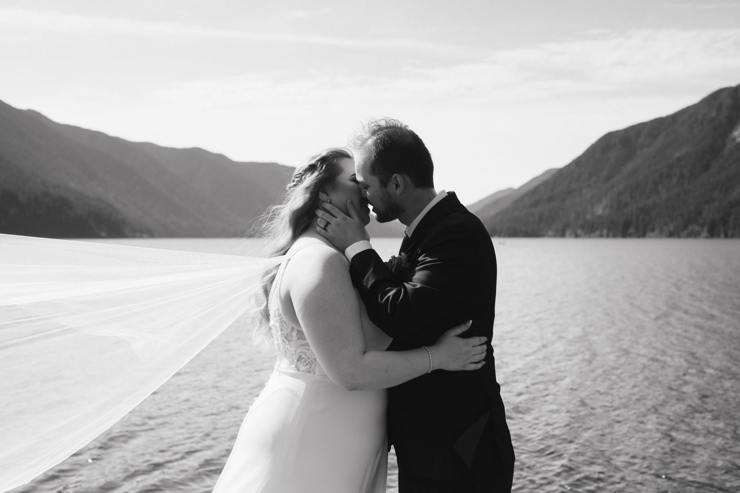 Black and white wedding photo of a couple embracing by a serene mountain lake.