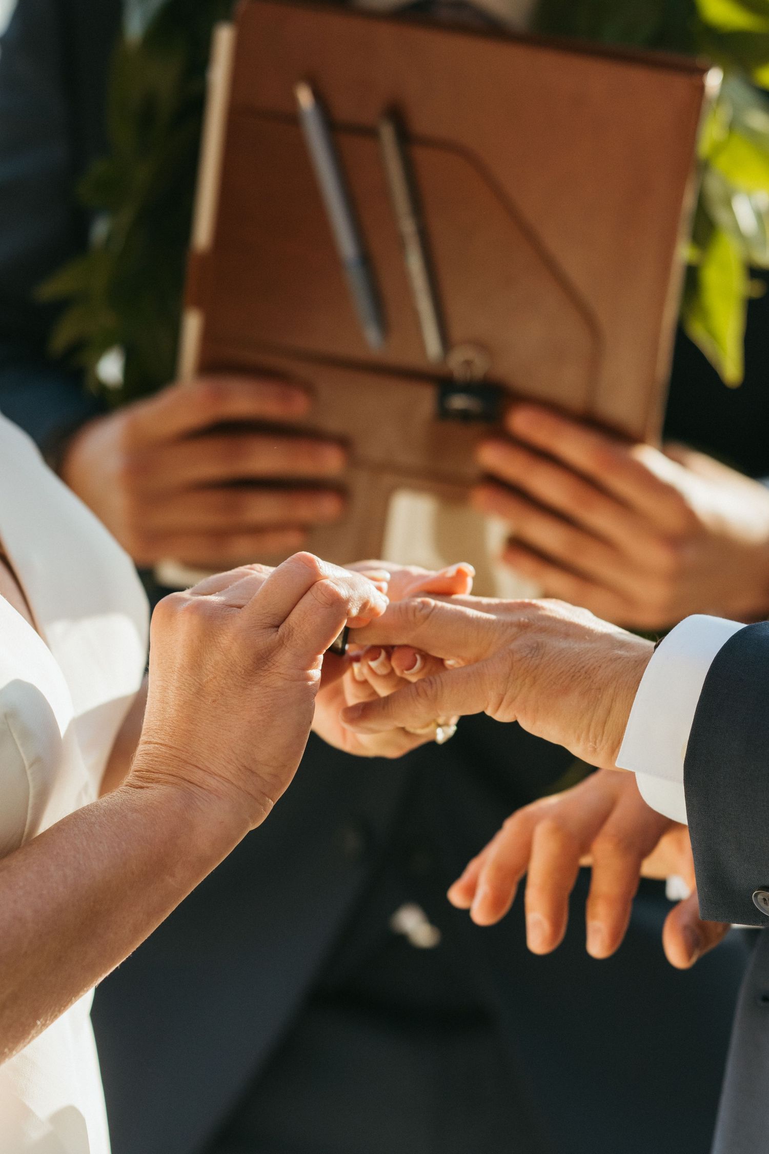 Close up of hands exchanging rings during wedding ceremony with brown officiant book visible.