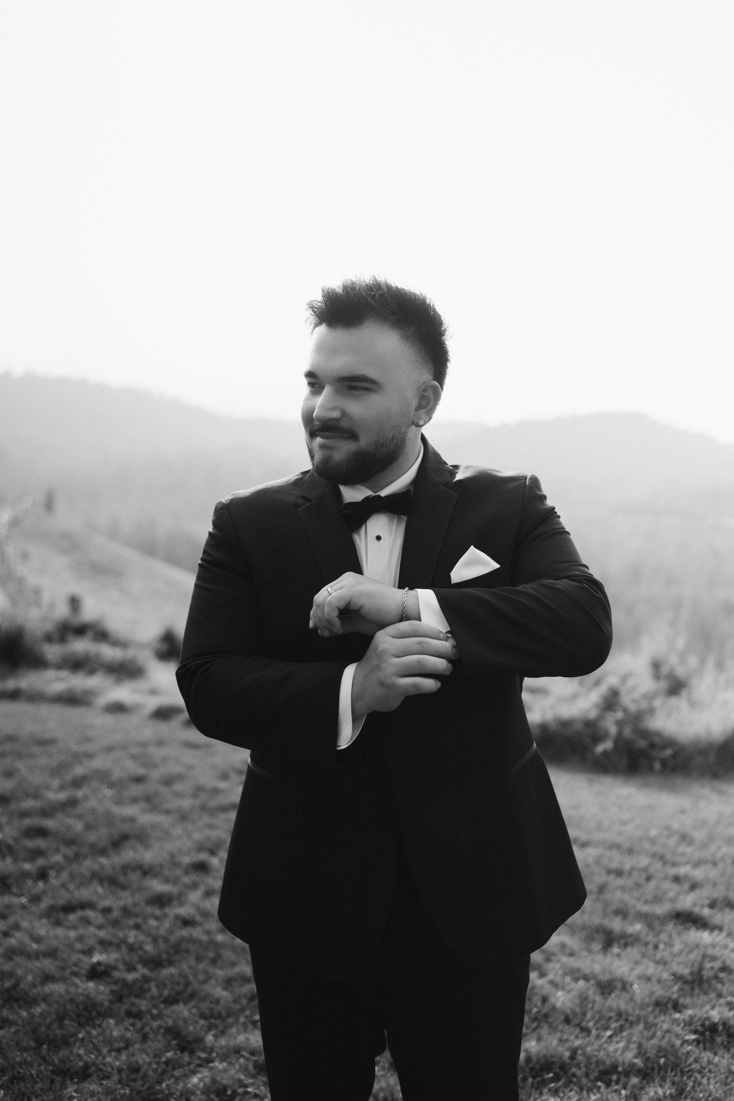 Groom in black tuxedo adjusts his cuff while standing on a hillside in black and white.