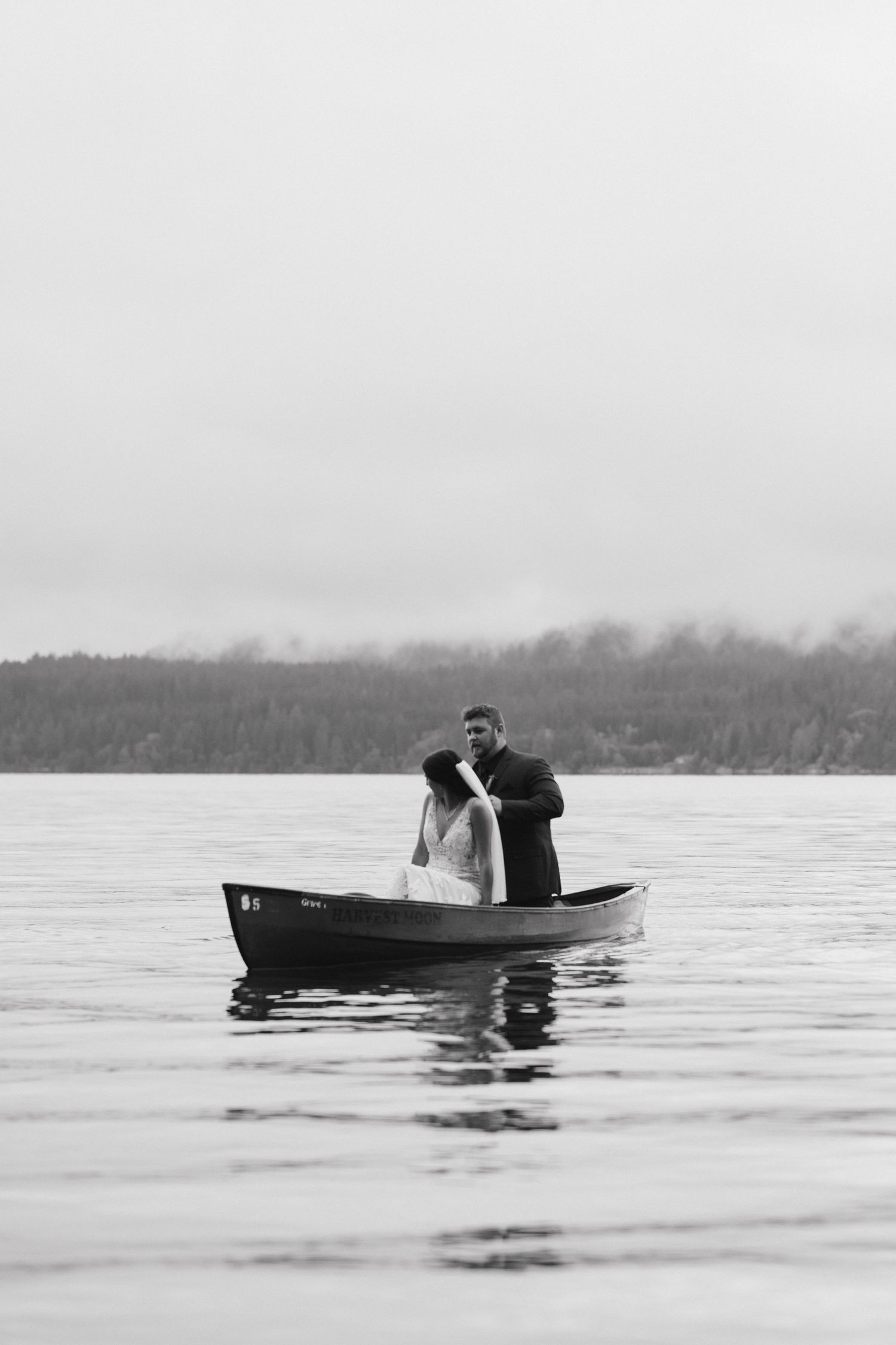 Misty black and white photo of a couple in a wooden boat on a serene lake surrounded by mountains.