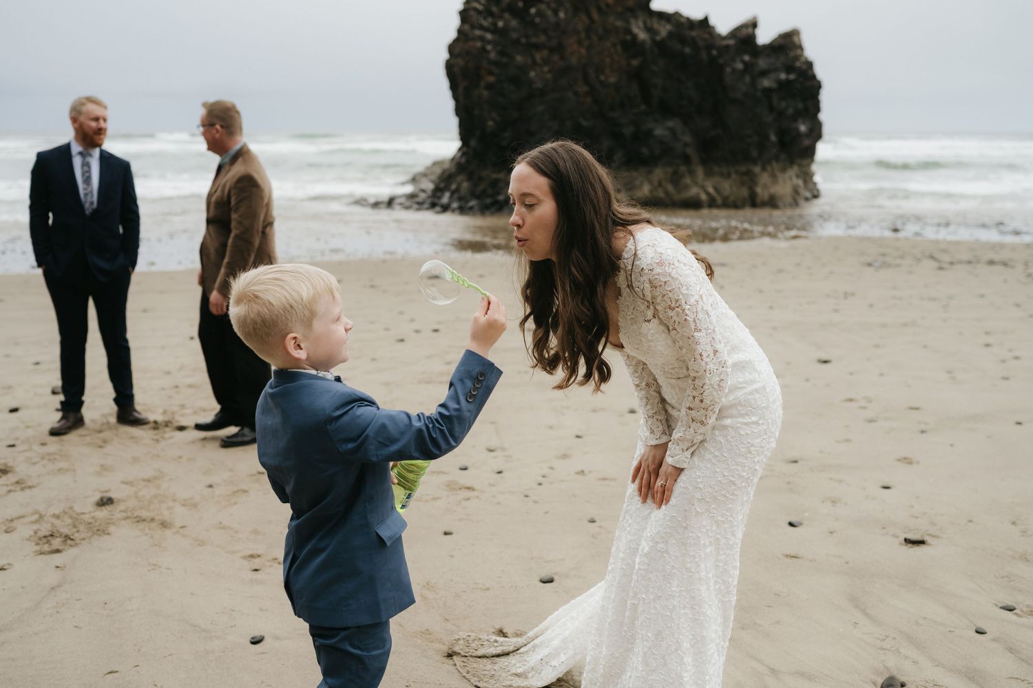 A bride in a lace dress bends down to interact with a young boy in a suit on a beach.