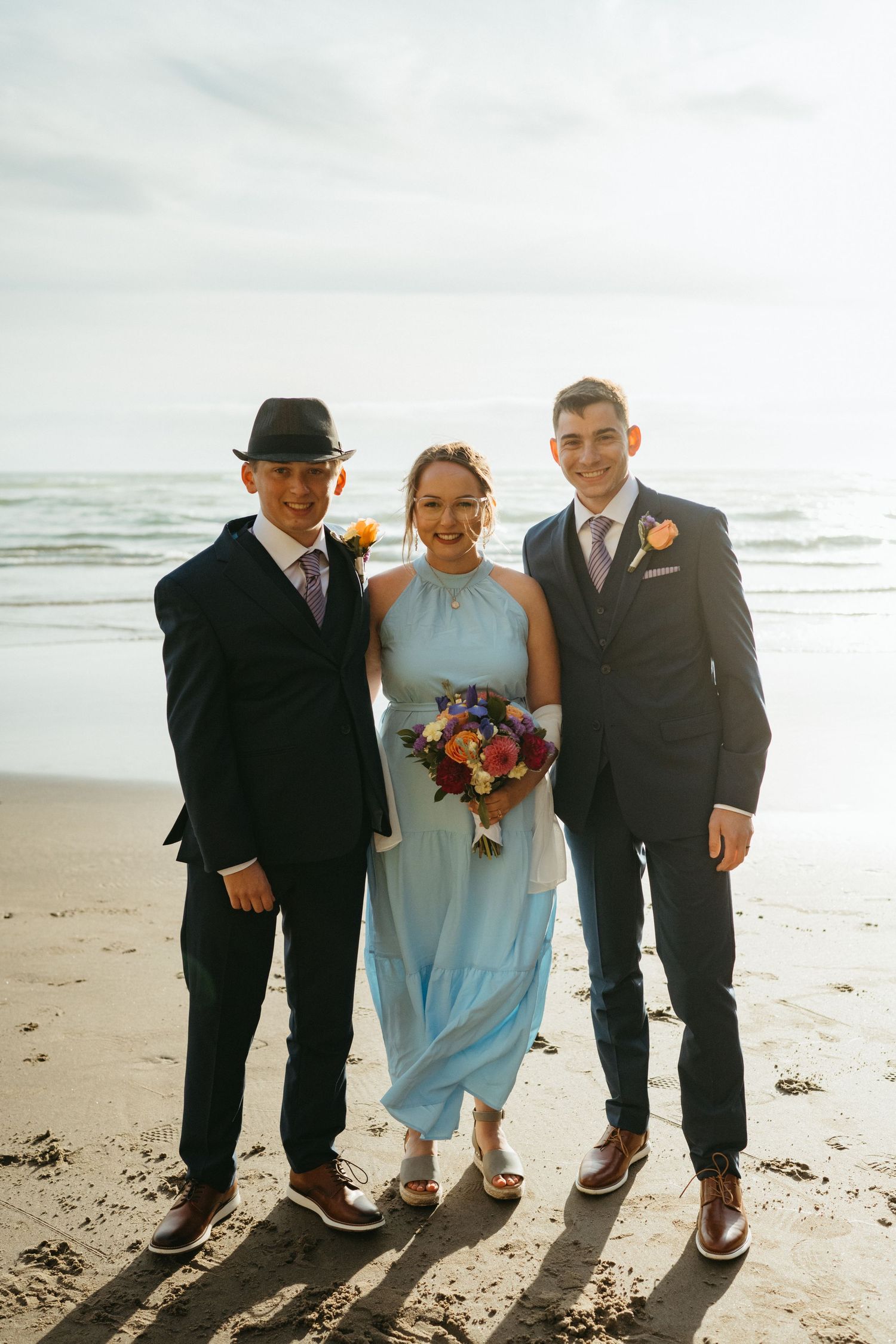 Three wedding attendants in formal wear pose on a beach at sunset.