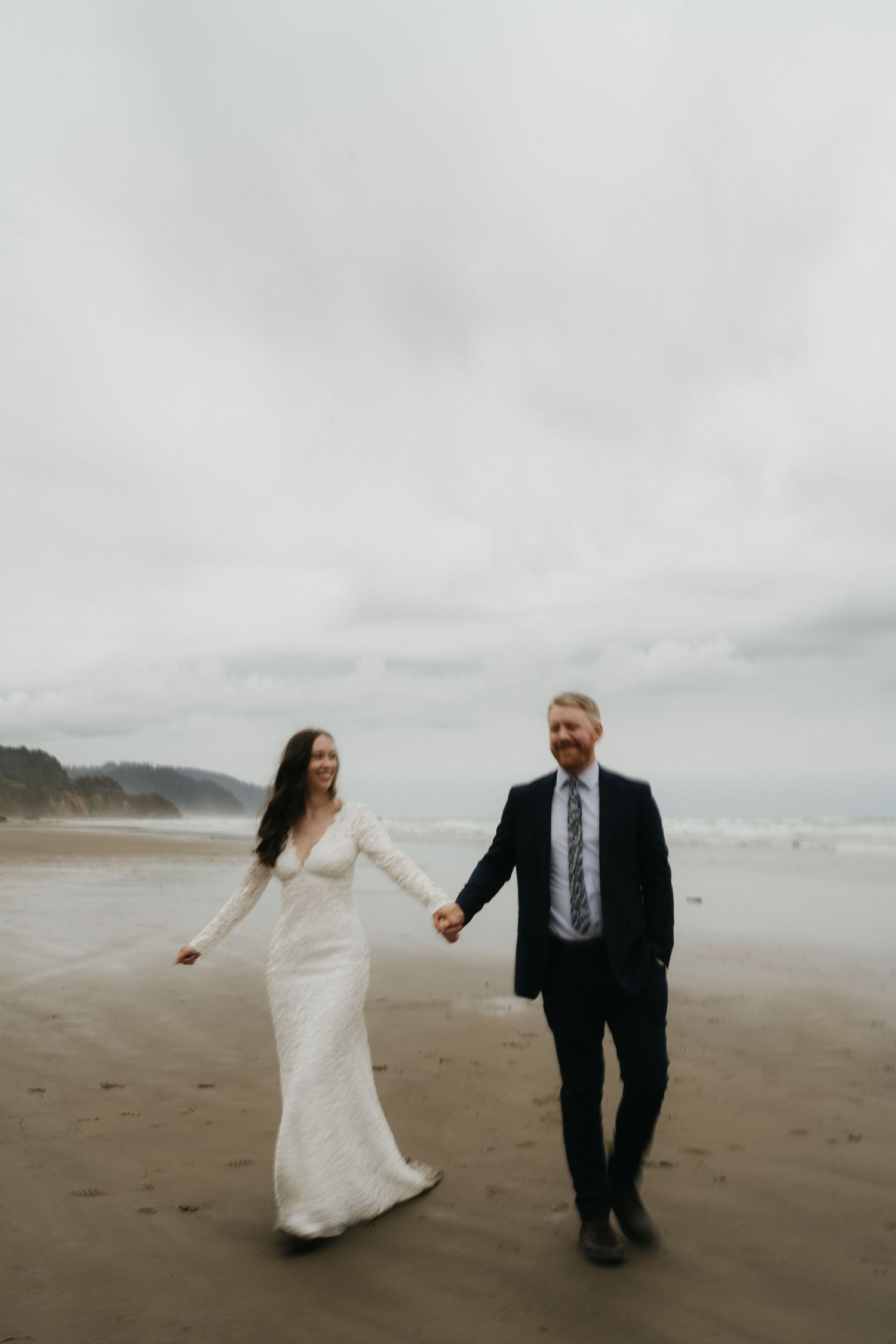 Newlyweds walking hand in hand along a scenic beach under overcast skies.