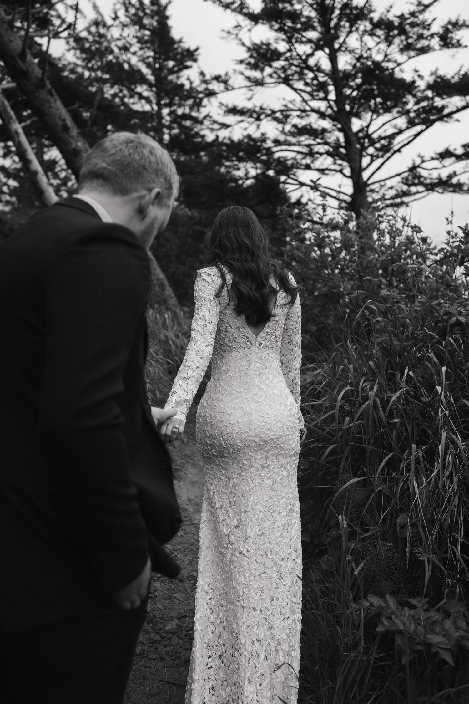 A black and white wedding portrait showing a couple among tall grass and pine trees.