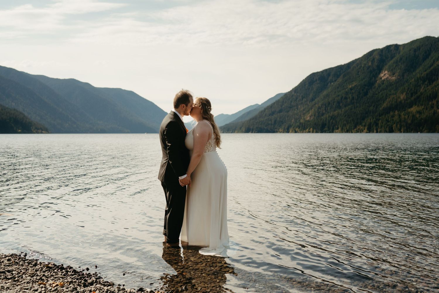 Newlyweds stand together in shallow lake water with mountains reflecting in the distance.