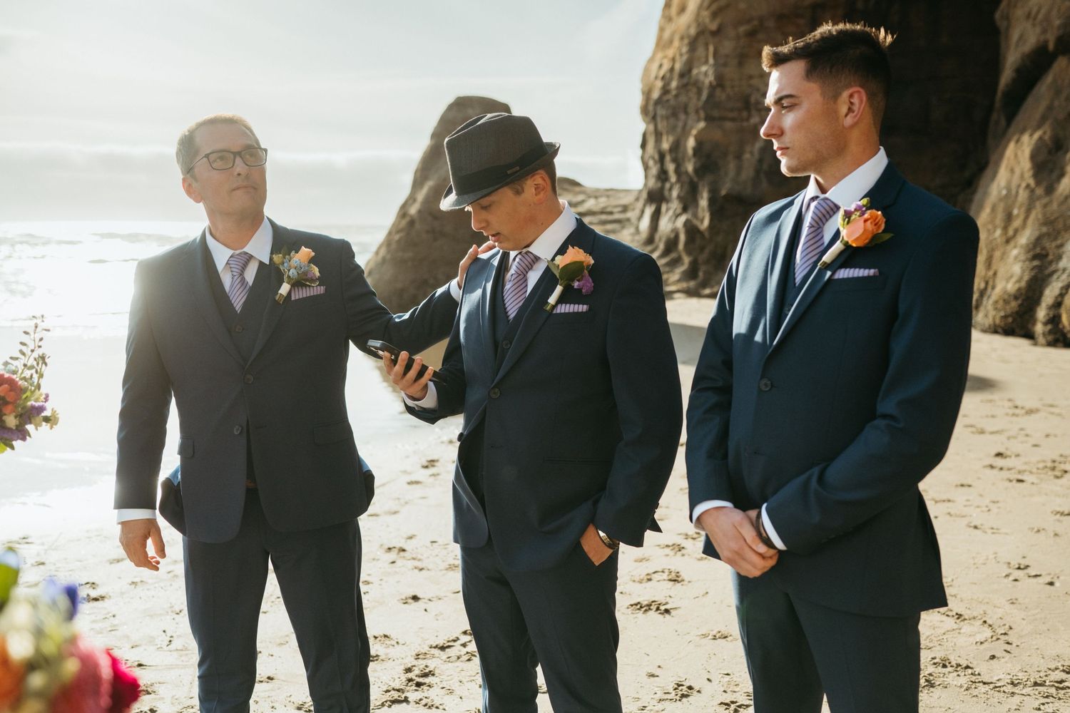 Groomsmen in navy suits share lighthearted moment on beach near cliffs.