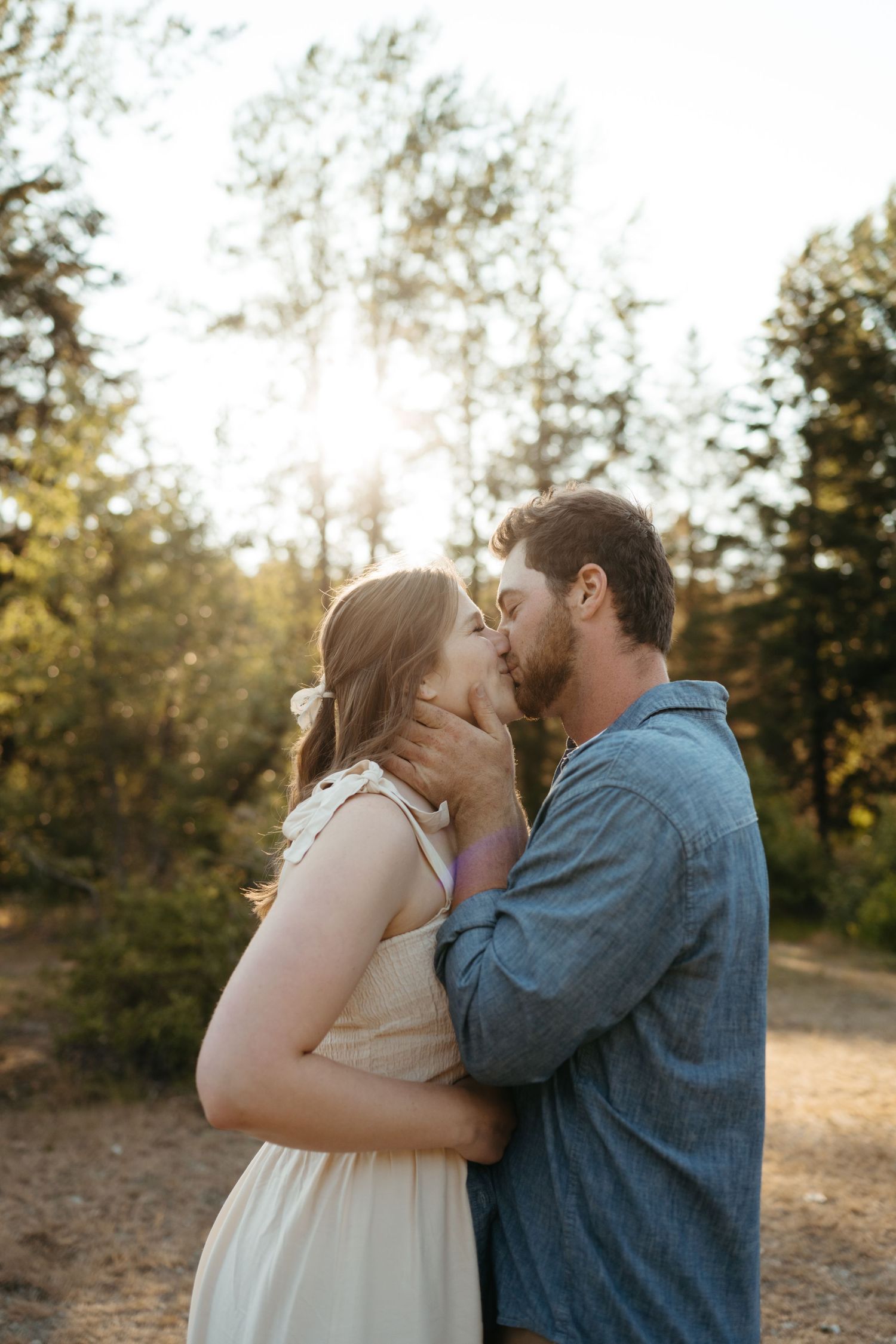 Romantic couple sharing an intimate kiss on a sunset forest trail, surrounded by evergreen trees.