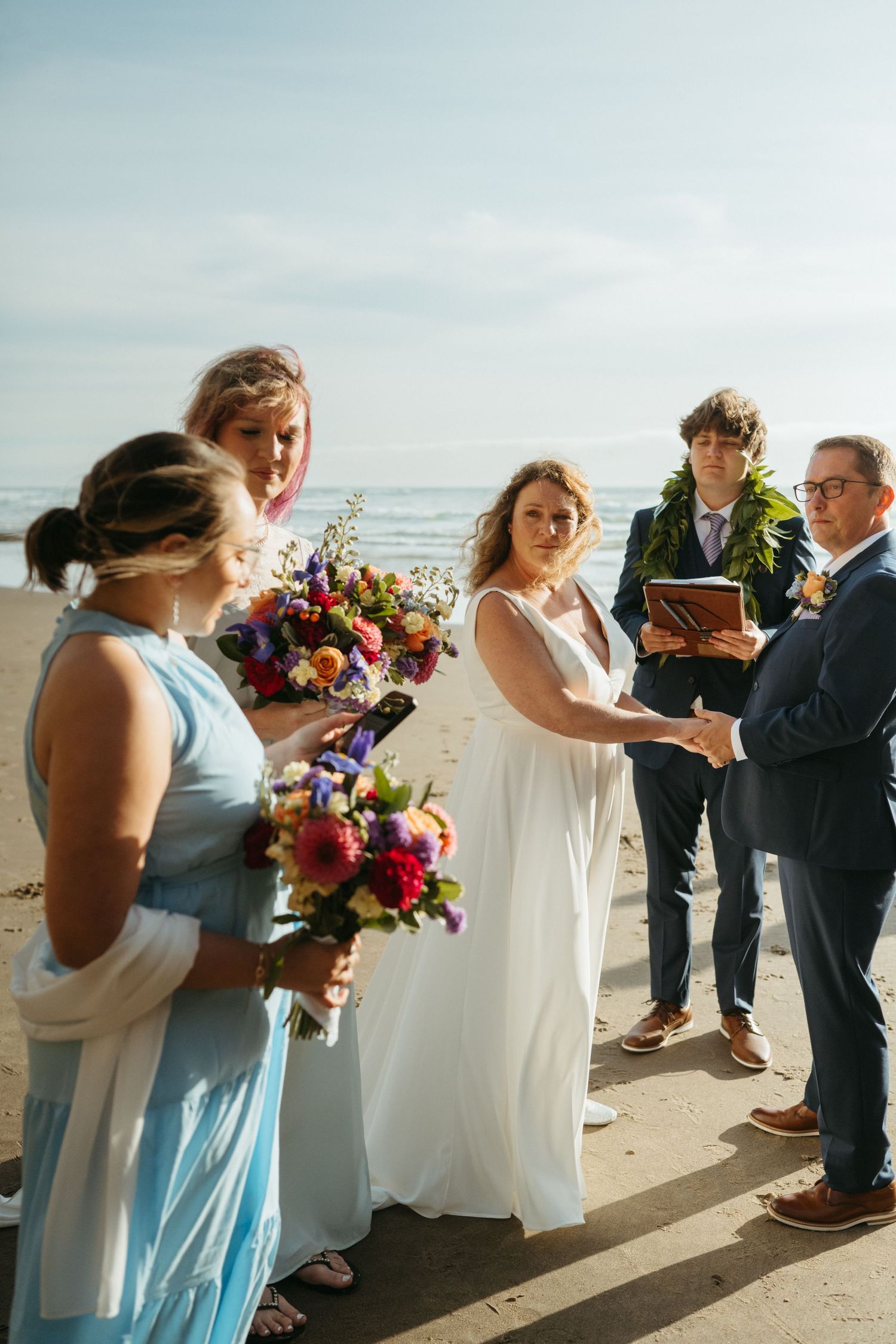 Beachside wedding ceremony with attendants holding colorful bouquets against ocean backdrop.