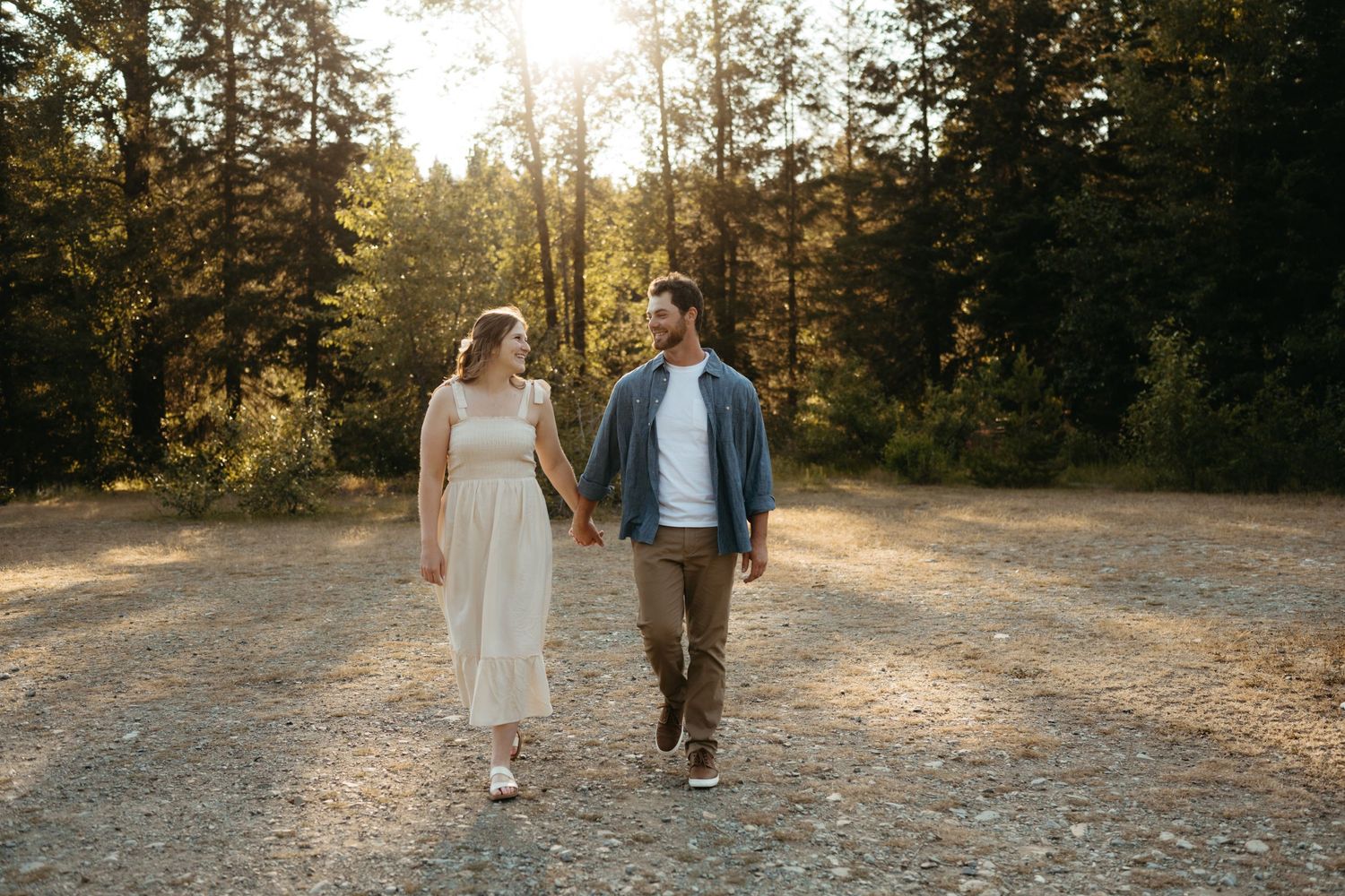 Couple walking hand in hand on a forest path during golden hour wearing casual attire.