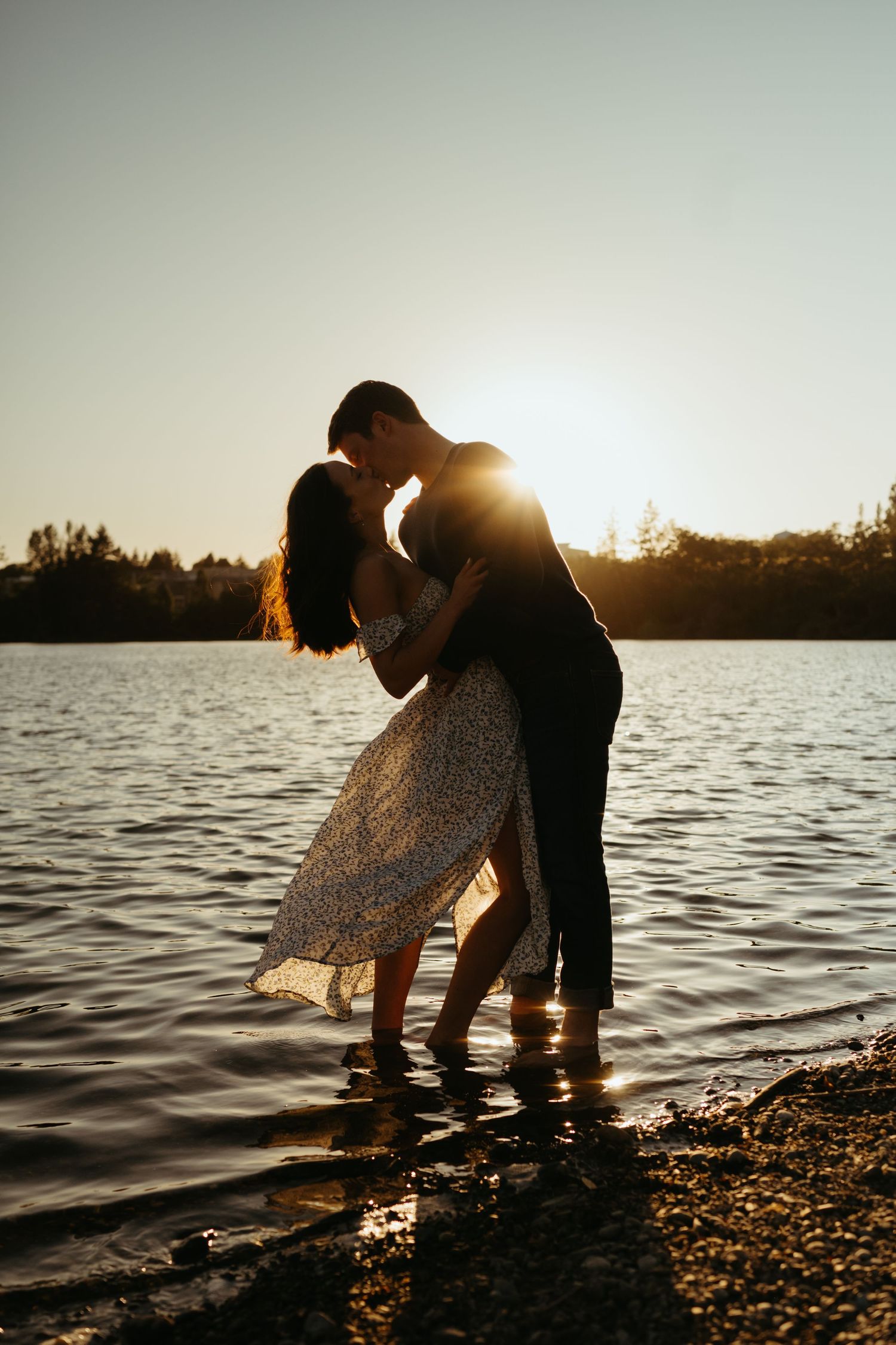 A romantic silhouette of a couple dancing in shallow water during sunset by a lake.