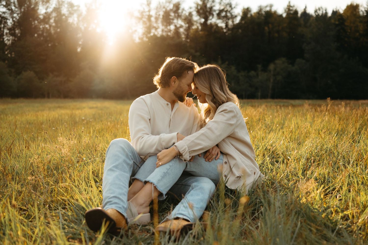 A couple in matching cream sweaters sits intimately in a sunlit meadow at sunset.