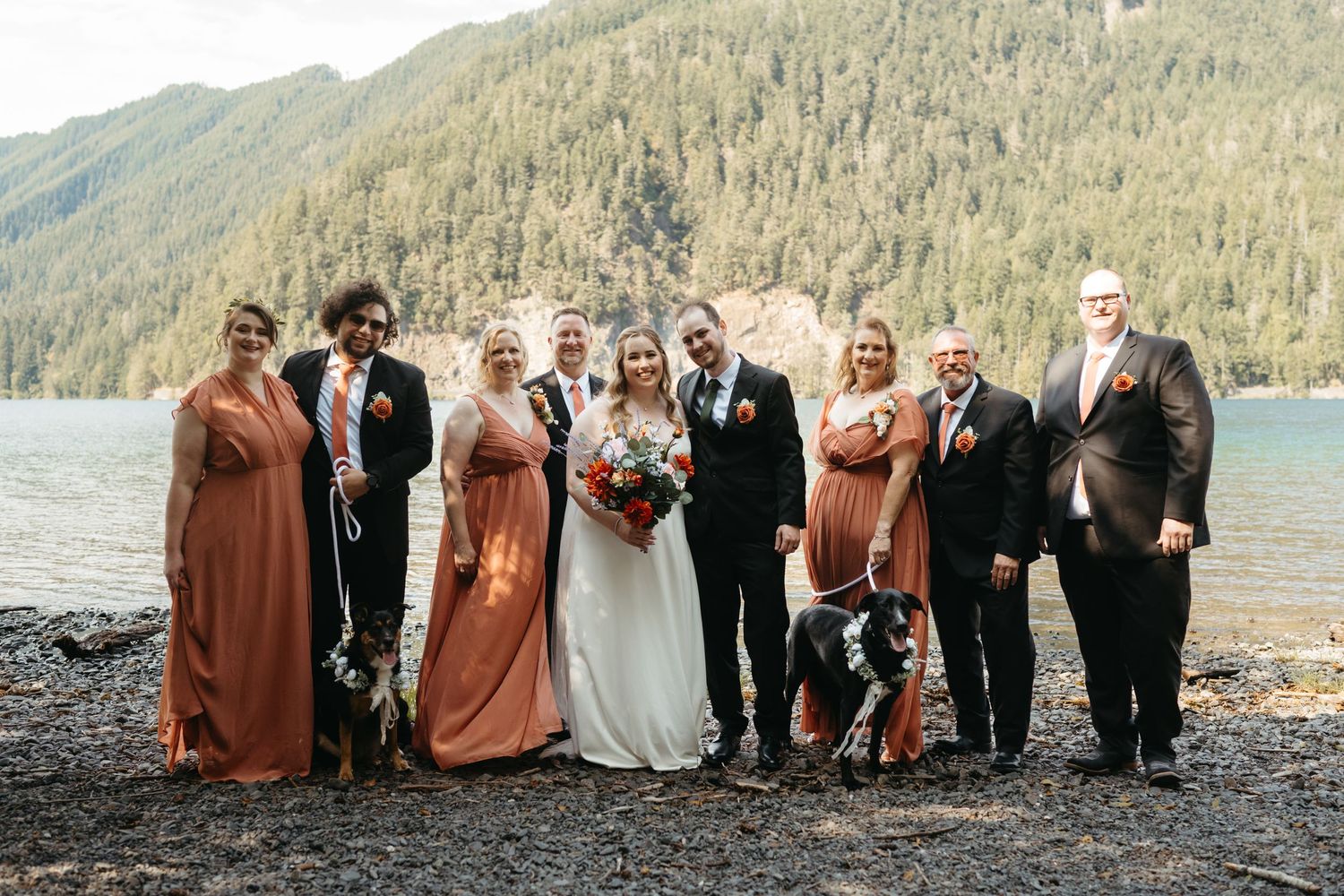 Wedding party in orange and black attire posing with dogs by a mountain lake.