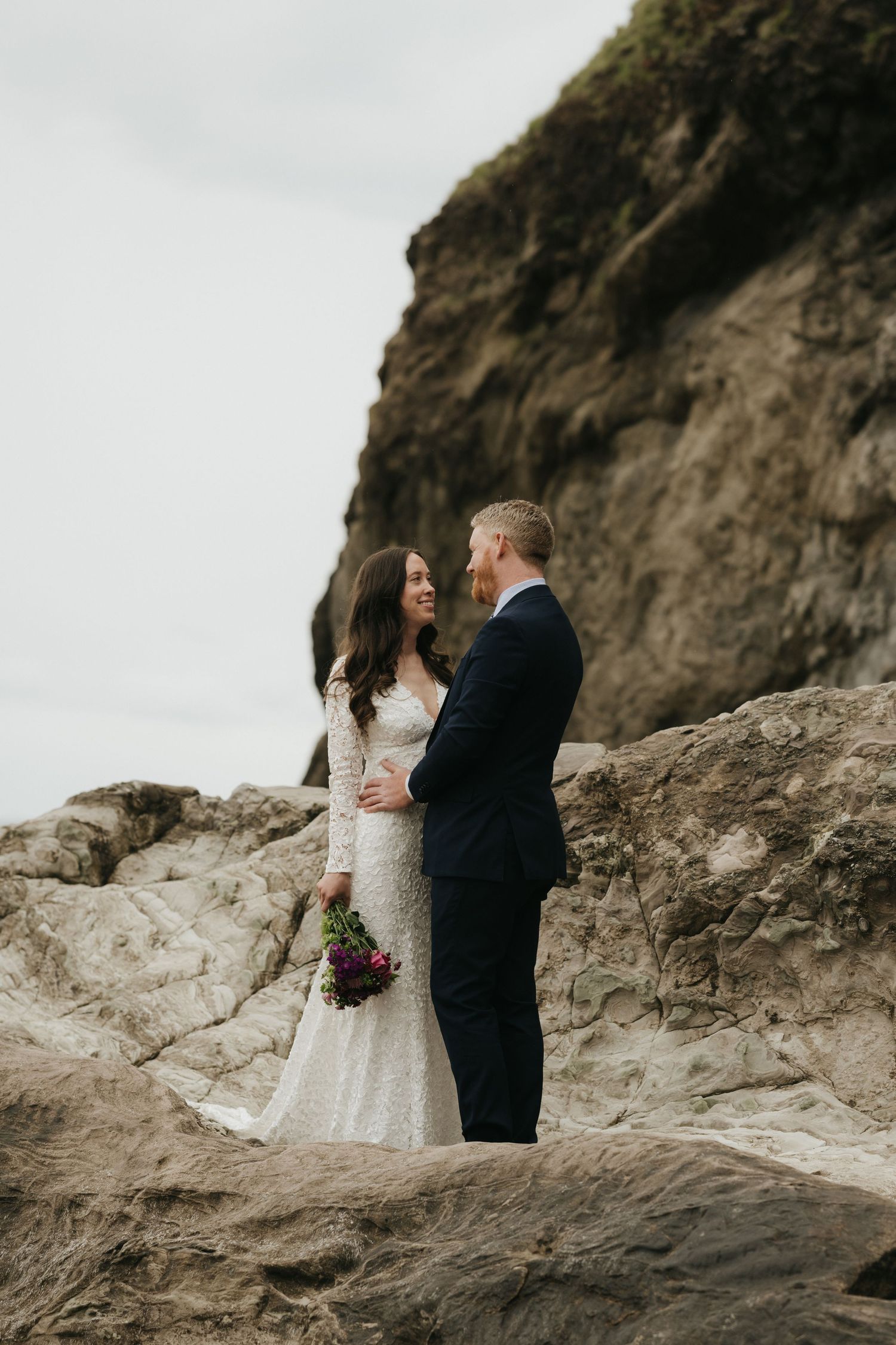 Intimate moment between bride and groom on rocky outcrop with coastal cliff behind them.