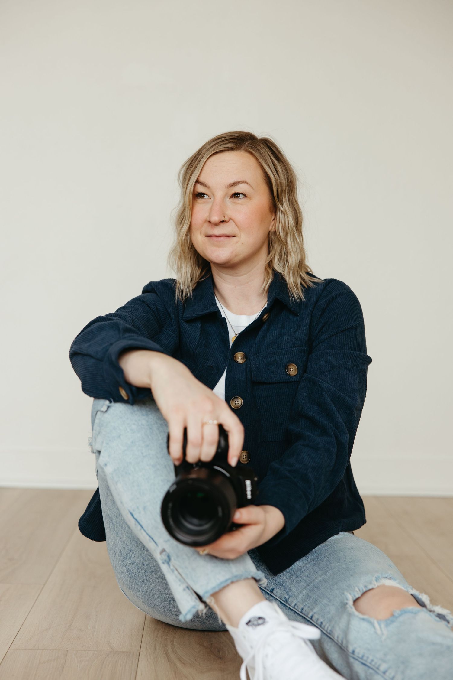 A blonde woman sits on a wooden floor holding a camera, wearing a denim jacket and ripped jeans, smiling thoughtfully.