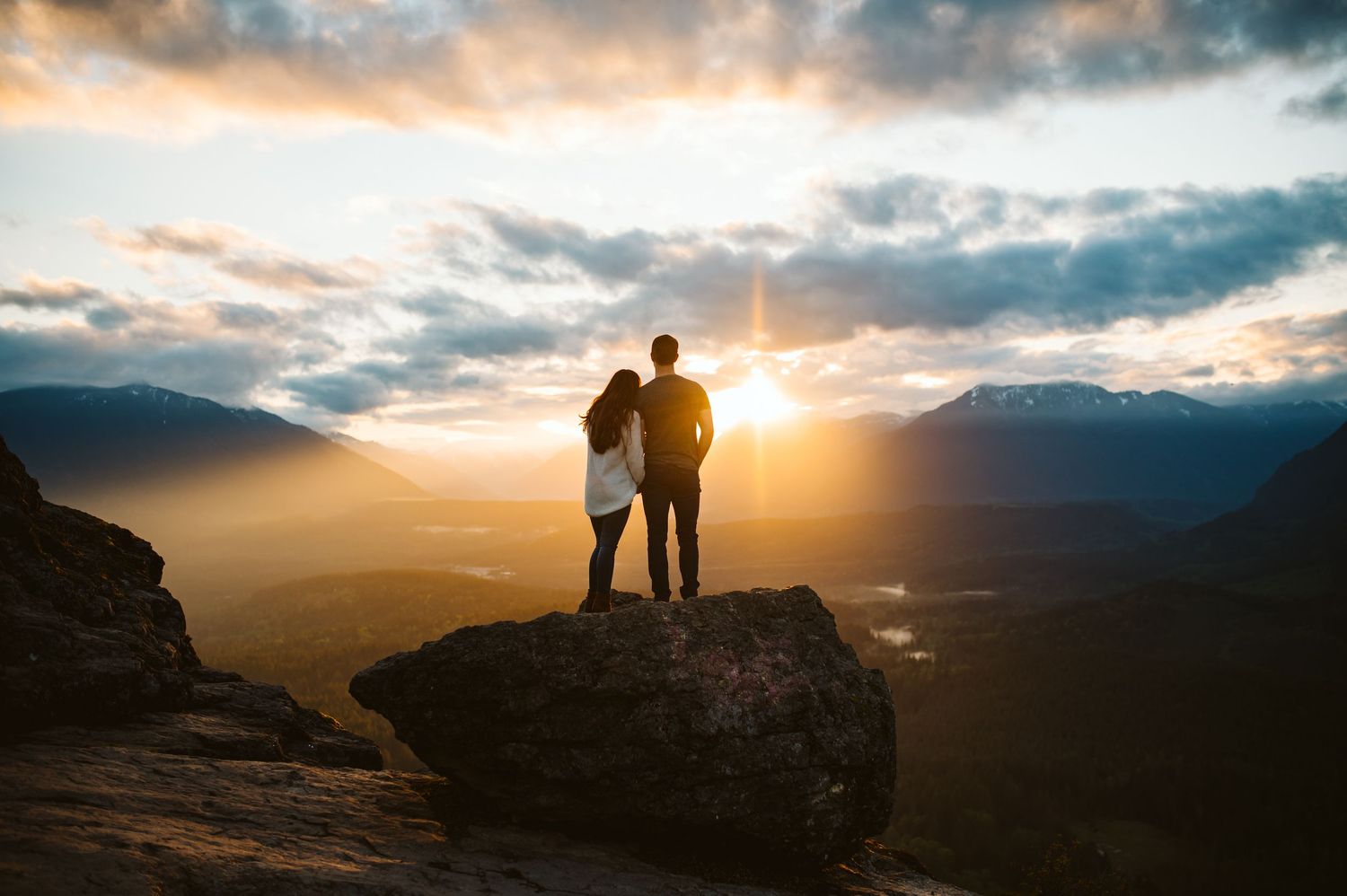 Two silhouettes stand together on a rocky overlook during a dramatic mountain sunset.