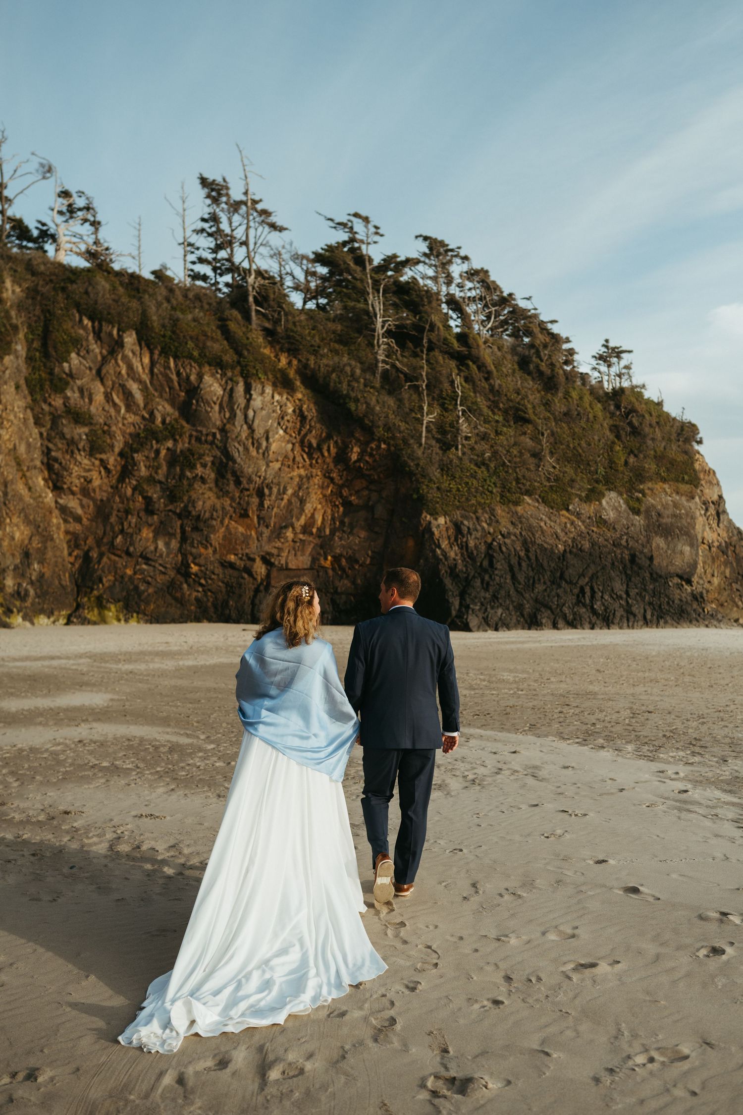 A couple walks hand in hand along a beach with dramatic cliffs in the background.