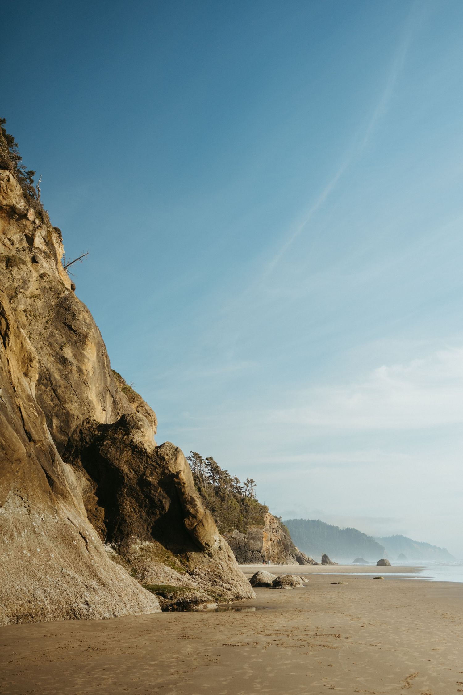Dramatic coastal cliff face meets sandy beach under clear blue sky.