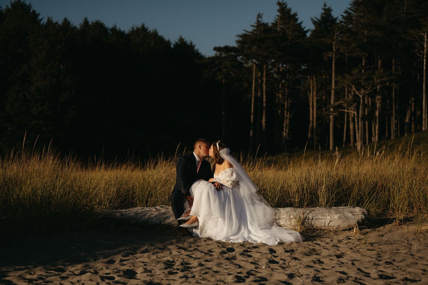 Couple in wedding attire sitting on rocks at sandy beach near forest at dusk.