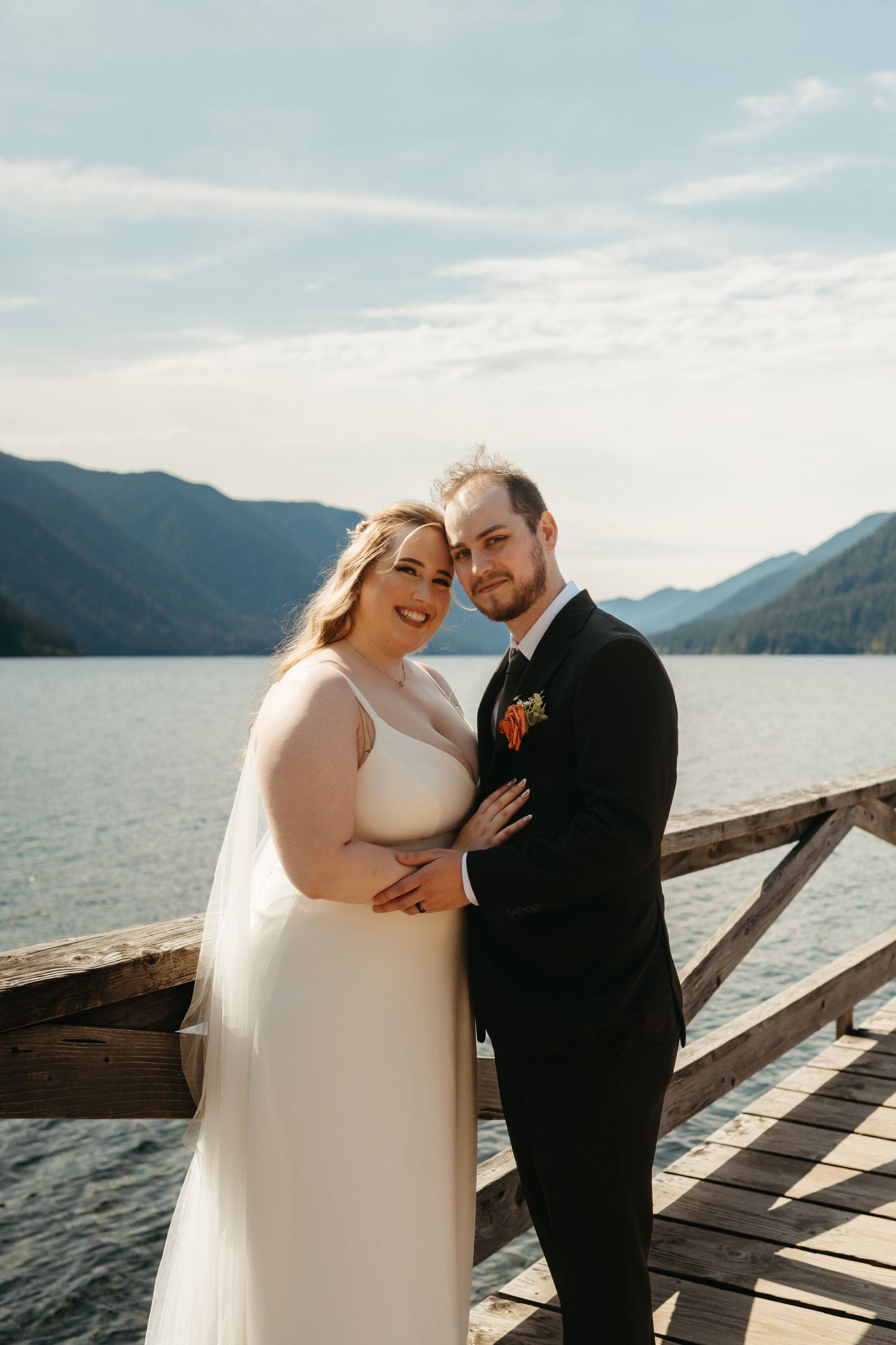 Wedding couple in elegant attire share a tender moment on a lakeside dock with mountain views.