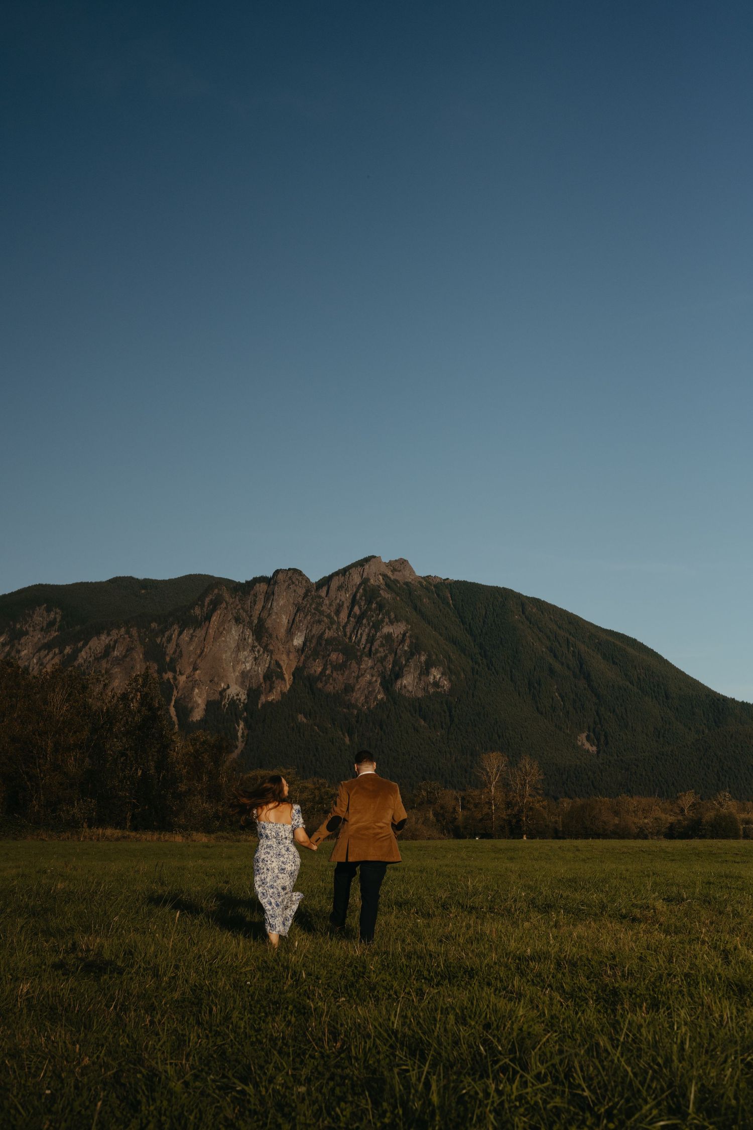 Couple walks hand in hand through grassy field with majestic mountain silhouette in background at dusk.