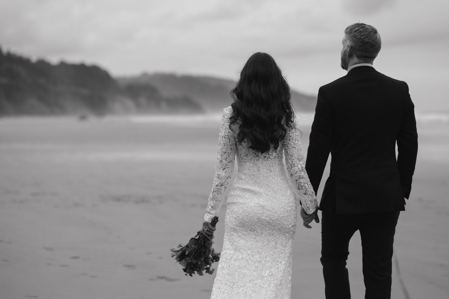 A wedding couple holding hands while walking on a misty beach in black and white.