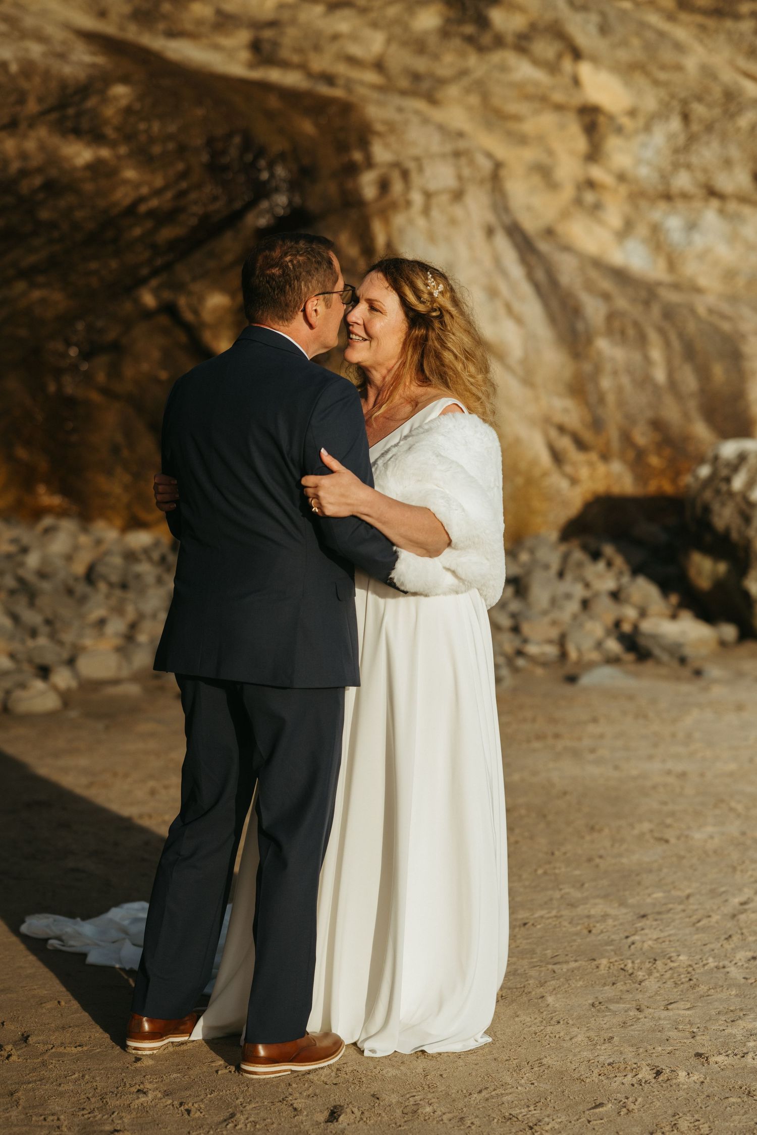 Couple embracing on beach with dramatic coastal cliffs in background.