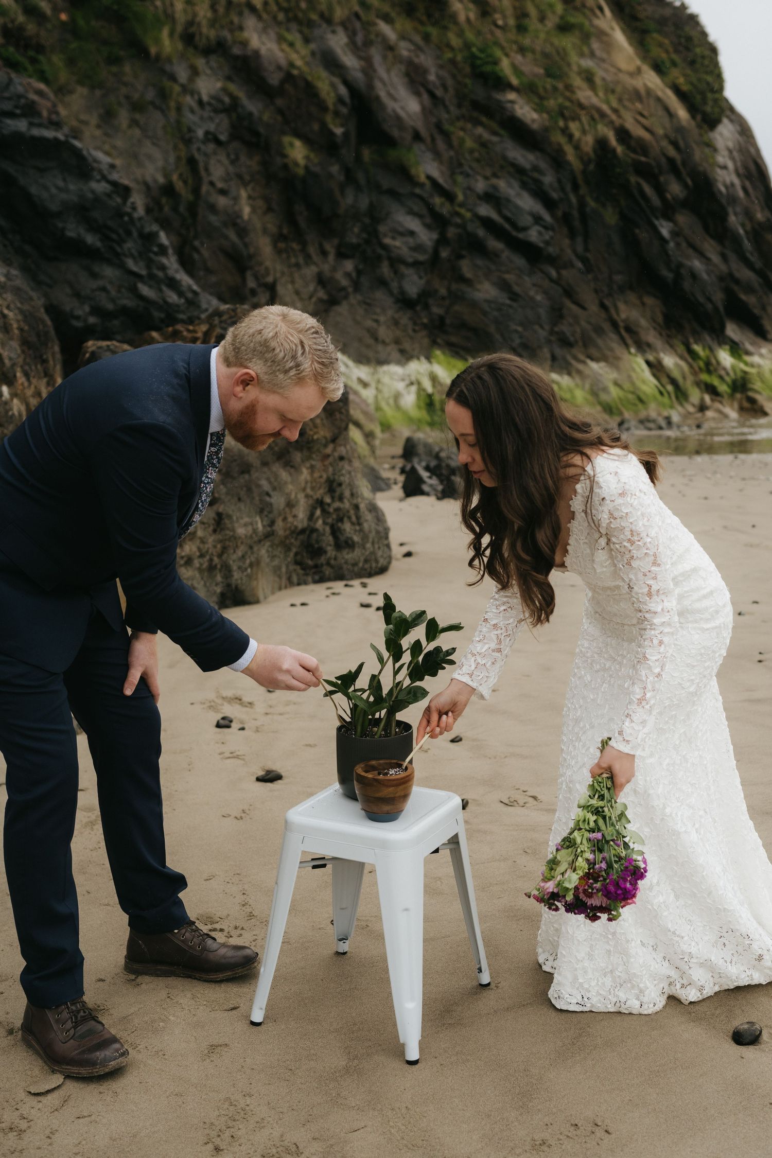 A couple arranges flowers on a white stool during their beach wedding ceremony.