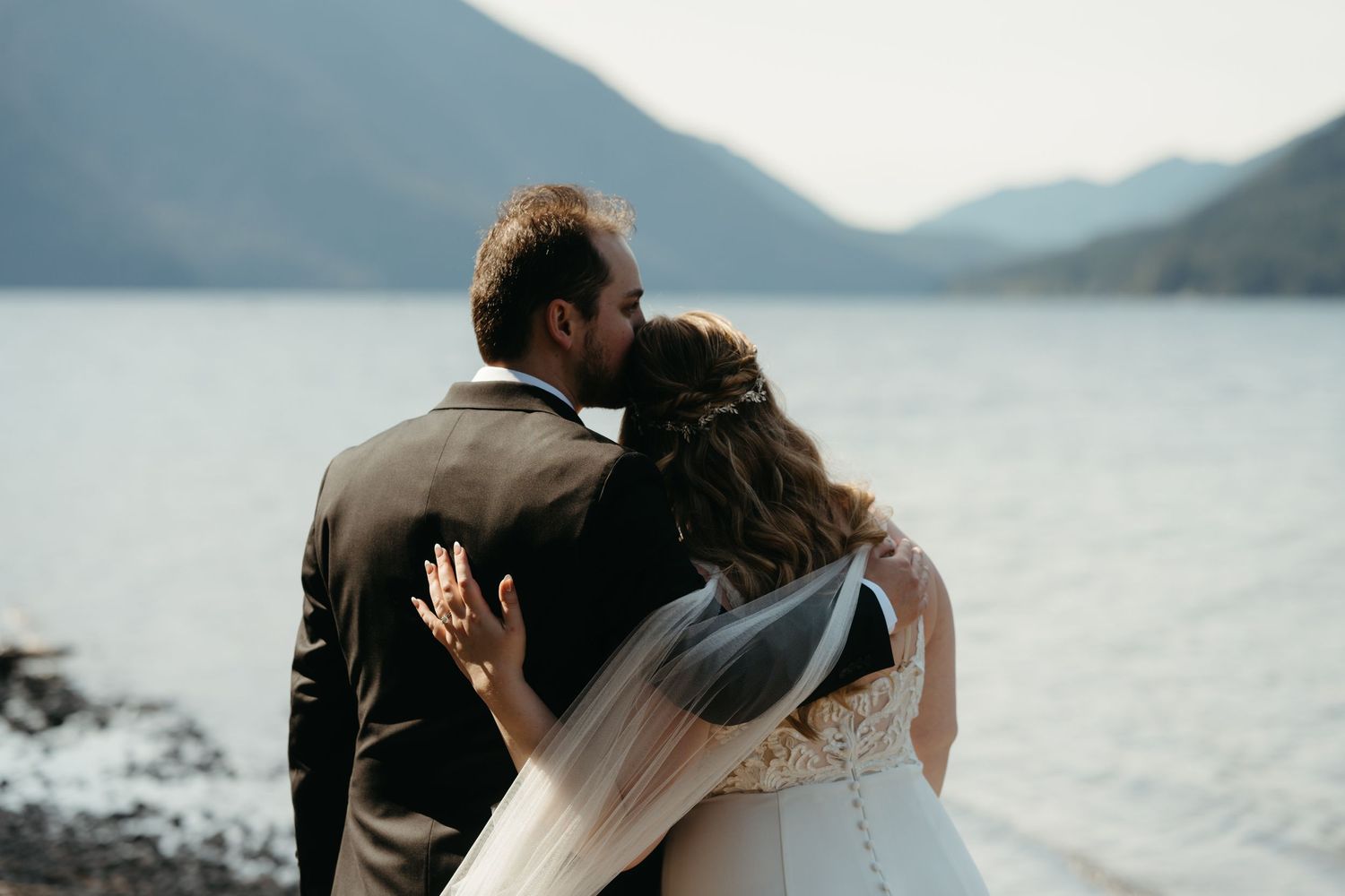 A couple embraces by a misty mountain lake, with the bride wearing a veil on their wedding day.