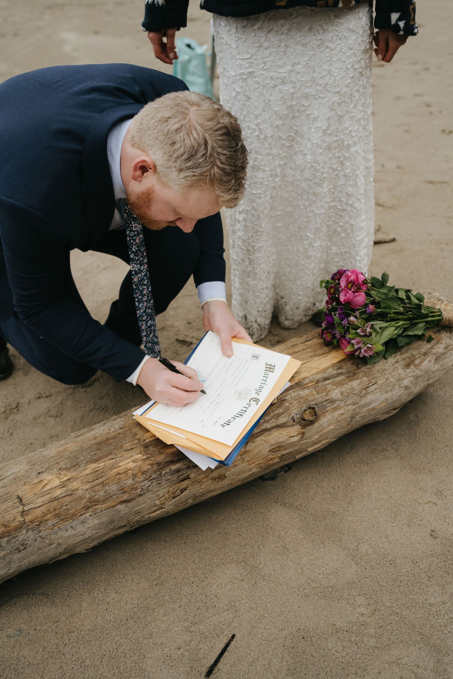 A person in formal attire signs papers on a piece of driftwood at the beach.