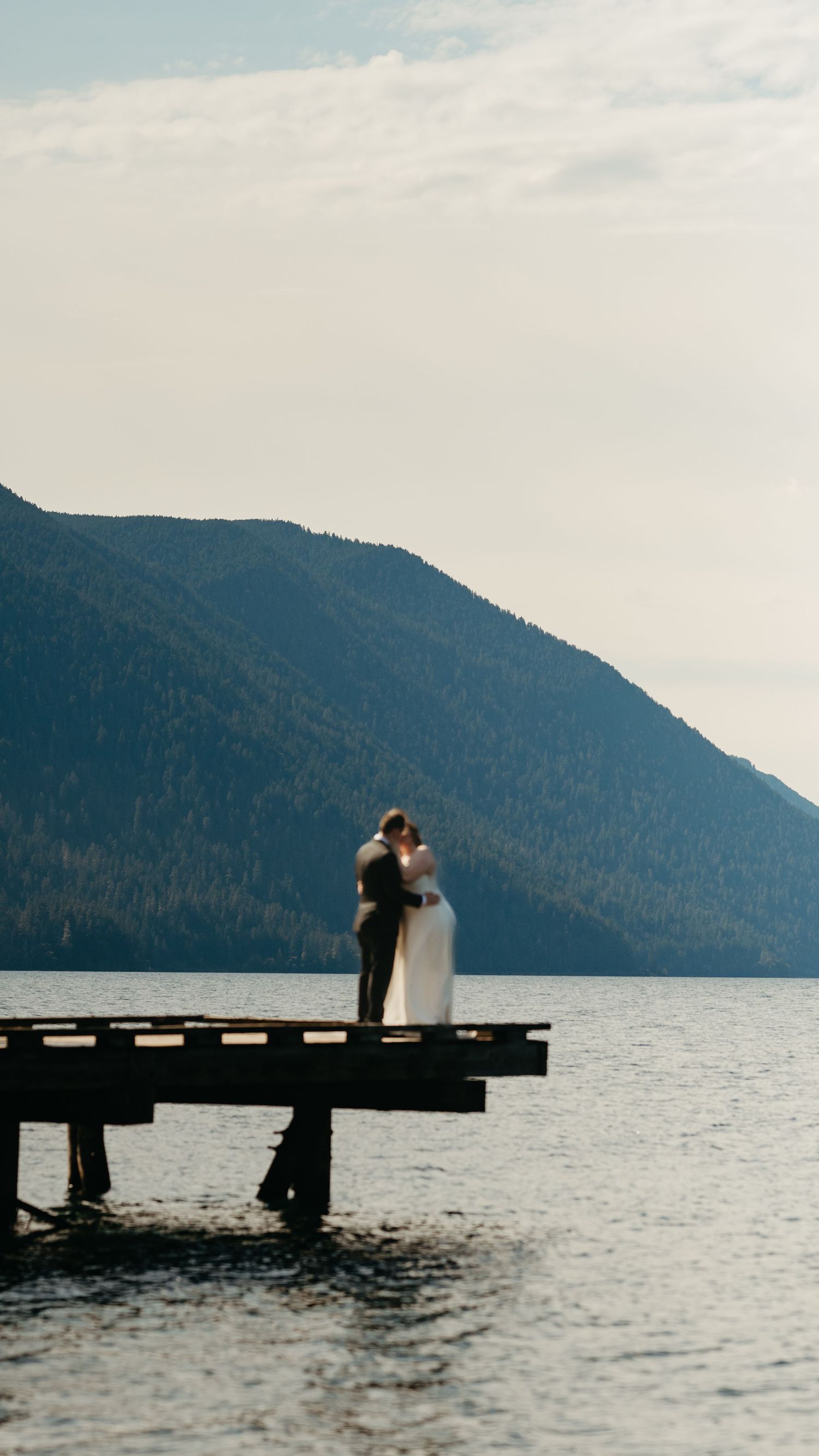 Romantic silhouette of newlyweds on dock with mountain lake backdrop during sunset.
