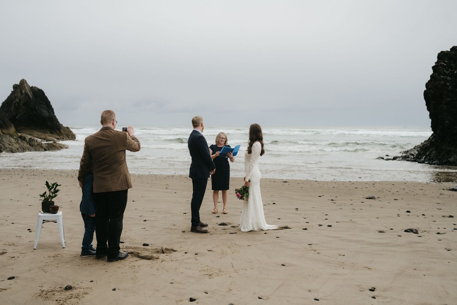 A small beach wedding ceremony taking place with dramatic coastal rocks in the background.