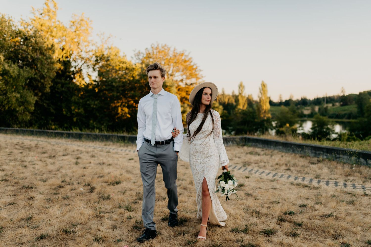 Couple walks hand-in-hand through golden field at sunset, bride in lace dress with hat holding bouquet, groom in gray suit.