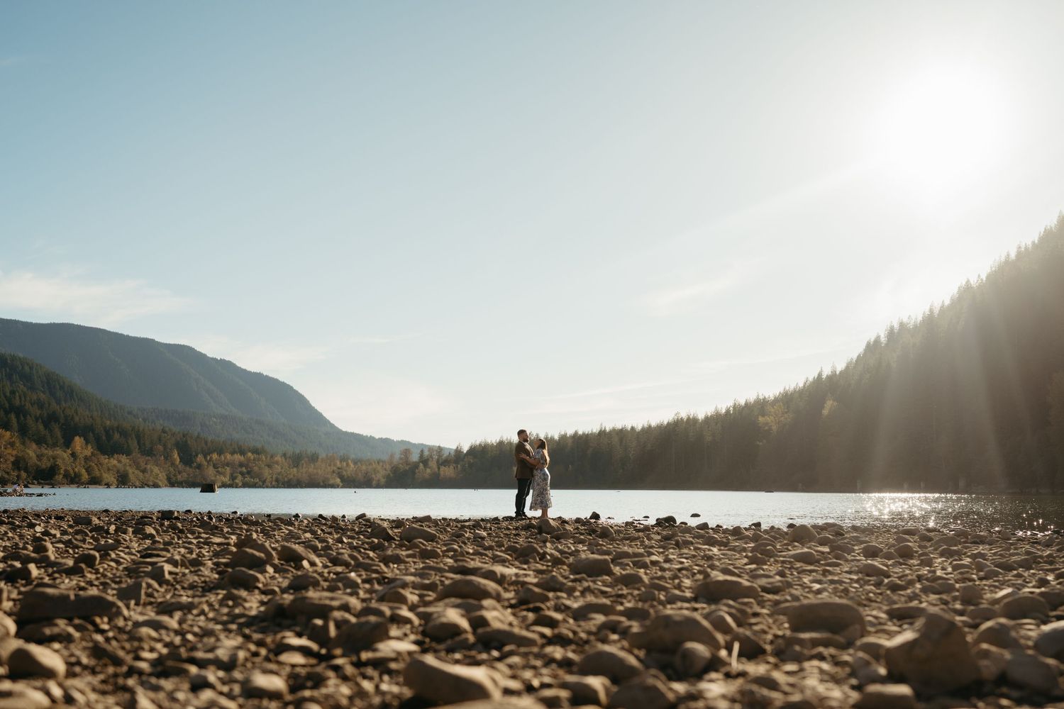 Two silhouettes stand together on a rocky shoreline with sunlit mountains and lake in the background.