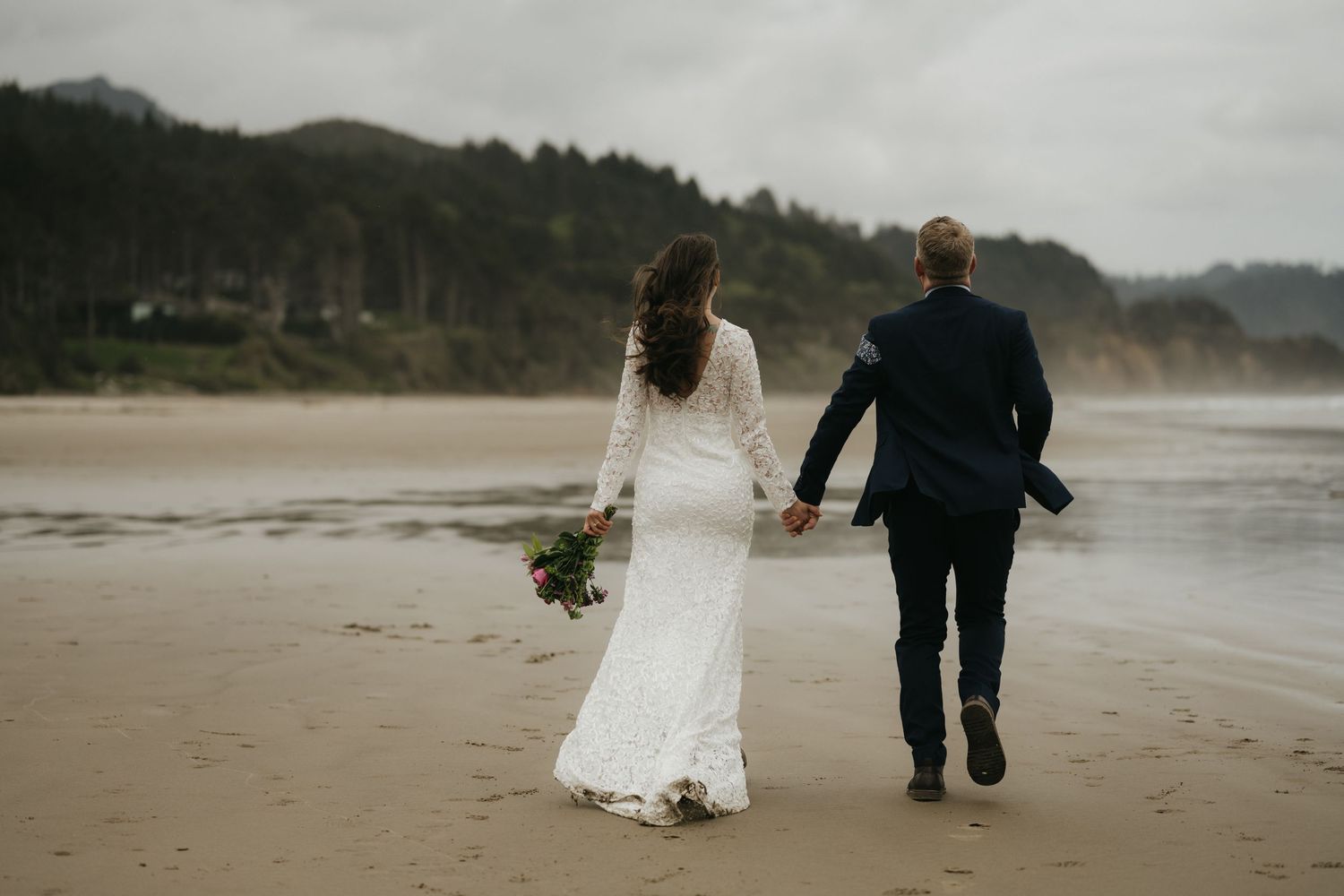 Wedding couple walking hand in hand on a misty beach with cliffs in the background.